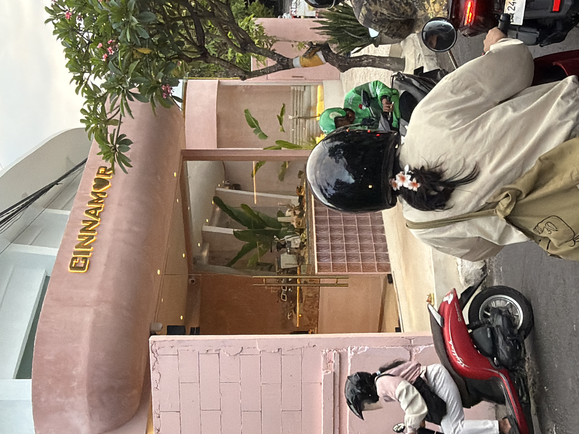This image captures a street scene in North Kuta, Indonesia, featuring a pink-colored building with the name "CINNOMAR" visible in gold lettering. The building appears to be a commercial establishment, possibly a cafe or shop, with a glass storefront displaying indoor plants and decor. In the foreground, several individuals are riding on motorcycles. One person in the foreground, viewed from behind, wears a black helmet and a light-colored jacket, with a beige bag slung over their shoulder. Another person on a motorcycle to their left wears a pink jacket and white pants, also wearing a black helmet. A third individual in a bright green jacket is visible on a motorcycle further back. The overall environment suggests a casual urban setting.