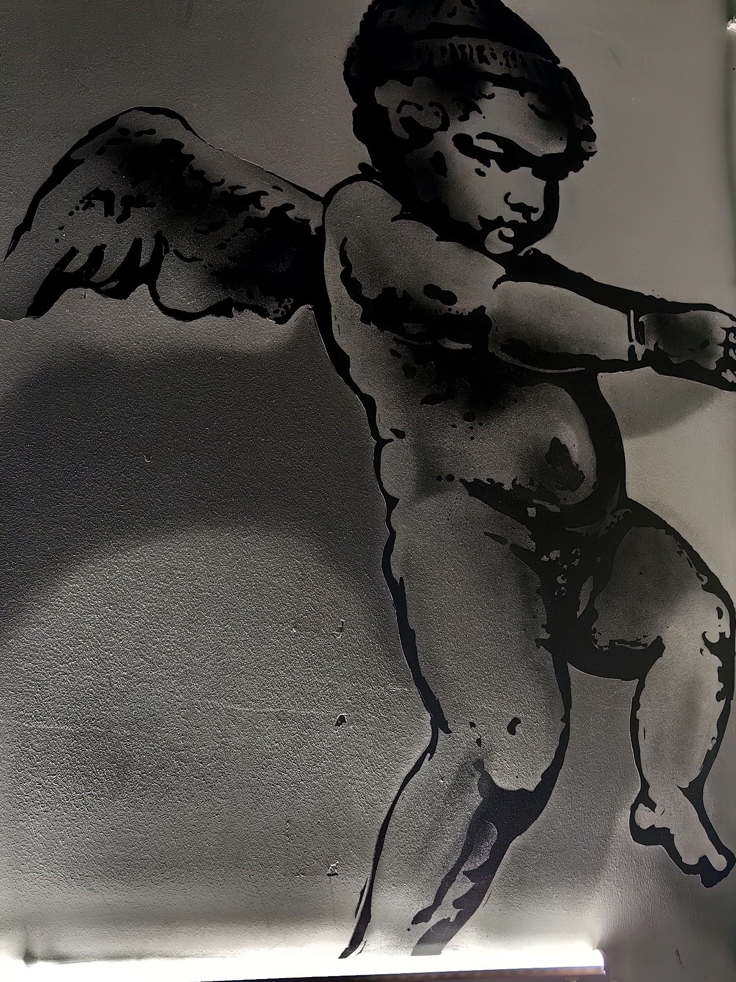 The image displays a stencil artwork of a cherub, rendered in black and grey tones against a textured, light-colored wall. The cherub is depicted in a dynamic pose, with its left arm extended forward and its right arm bent at the elbow. Its wings are spread wide, suggesting movement or flight. The cherub's facial features are clearly visible, with a downward gaze and a neutral expression. The overall style of the artwork is illustrative, with bold outlines and shaded areas creating a sense of depth and form. The wall's texture is granular and uneven, with some subtle shadows and highlights suggesting the lighting conditions. A bright light source is visible at the bottom of the image, casting a glow upwards. There is no visible text in the image. The artwork's style and subject matter evoke classical or Renaissance imagery.