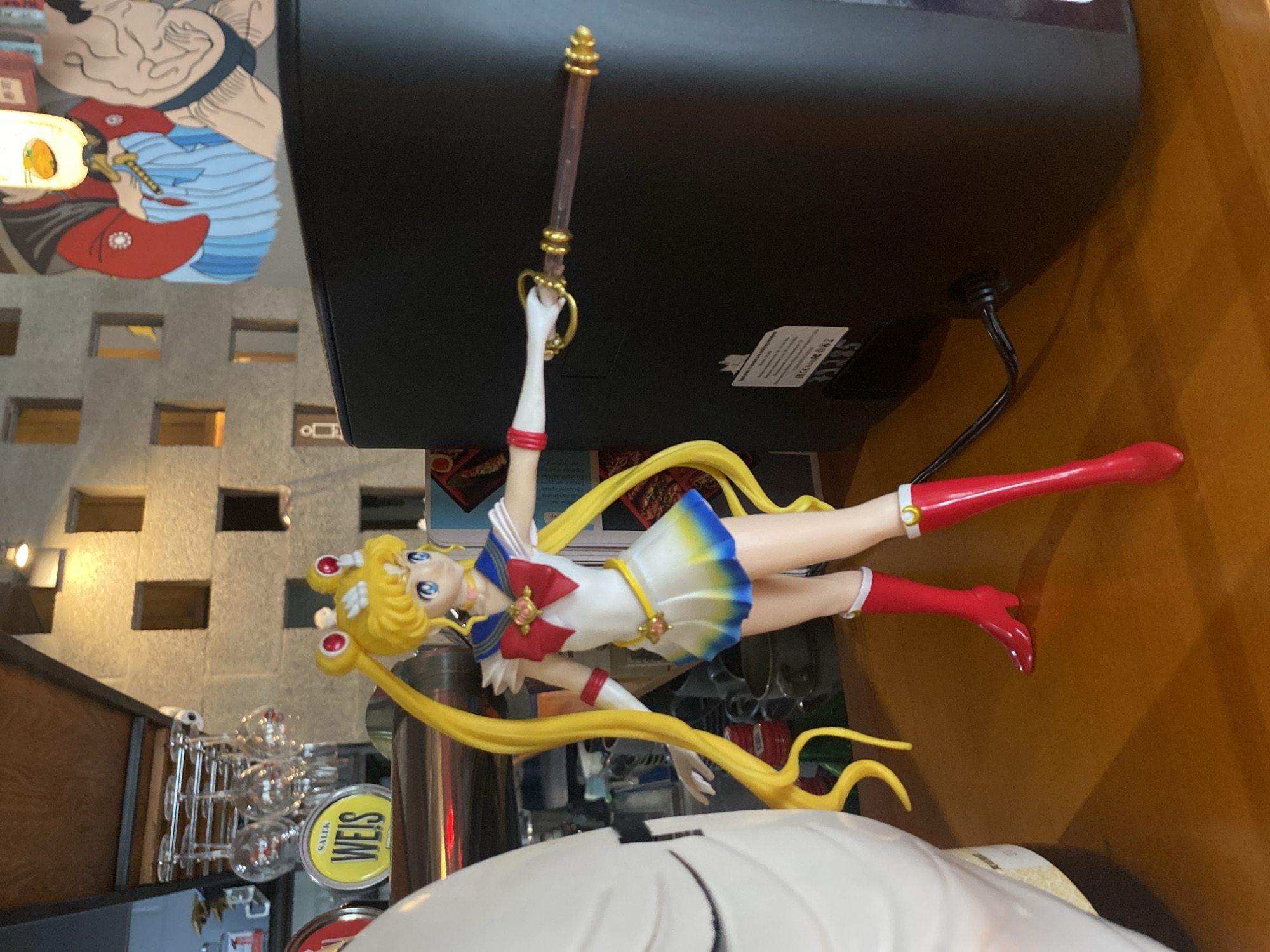 A Sailor Moon figurine is prominently displayed on a surface, with a black electronic device positioned behind it and to the right. A sword or wand is held aloft in the figurine's right hand. To the left and slightly behind the figurine is a wall with a textured, brick-like pattern. A series of clear drinking glasses hang in a rack below the wall. A "WEISS" logo on a yellow circle is partially visible near the glass rack. In the top left corner, a mural of a man is visible. The image was captured indoors. The location is Krasnoyarsk, Russia.