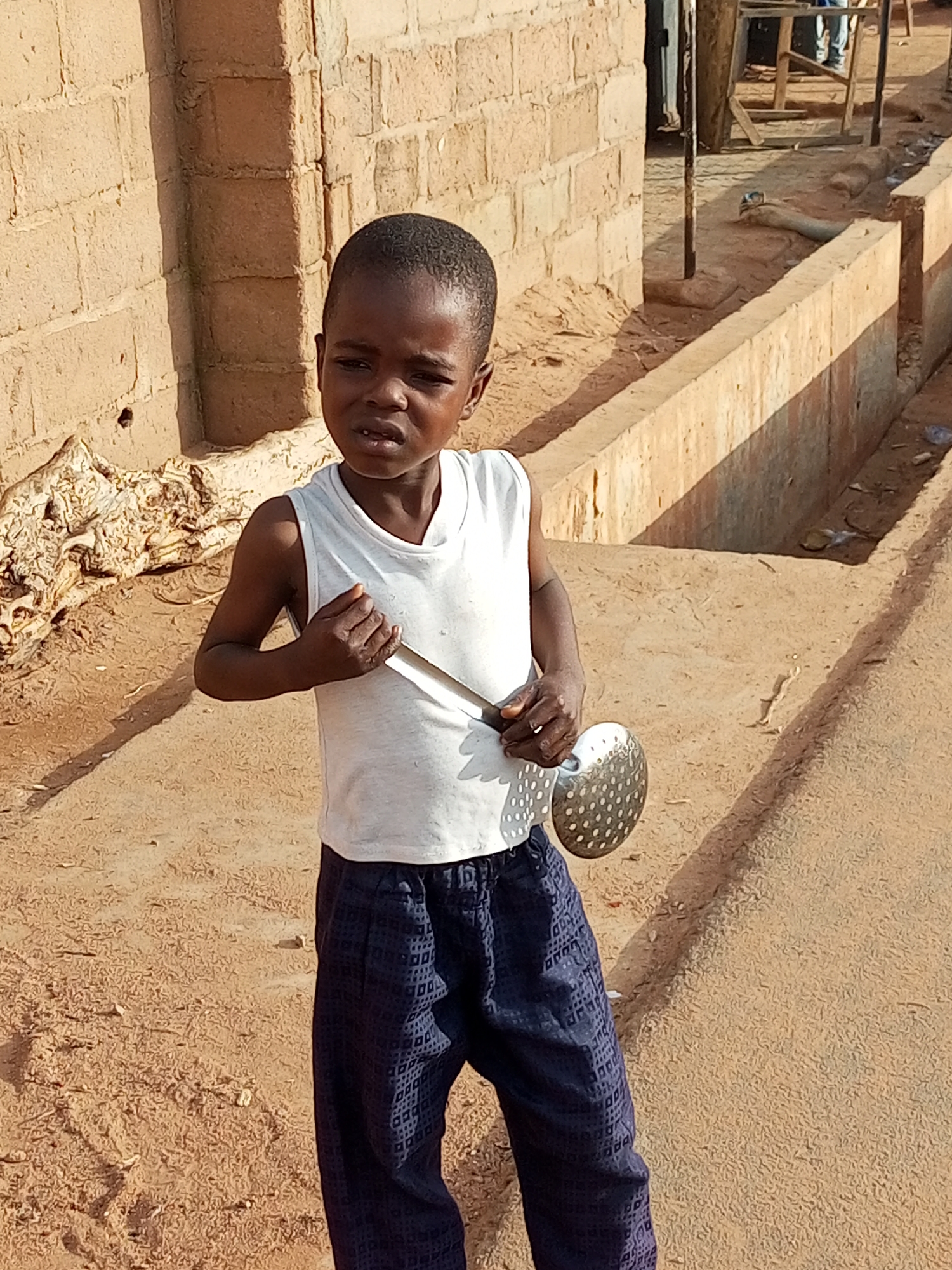 In the city of Unknown, Unknown, a young child stands outside near a brick building. The child, wearing a white sleeveless shirt and dark patterned pants, holds a metal skimmer with a perforated surface. The ground is sandy and dusty, with a shallow concrete channel running alongside. A pile of debris or dry vegetation is visible to the left of the child. In the background, wooden structures and more buildings can be seen. The overall scene suggests an outdoor, possibly rural or semi-urban environment.