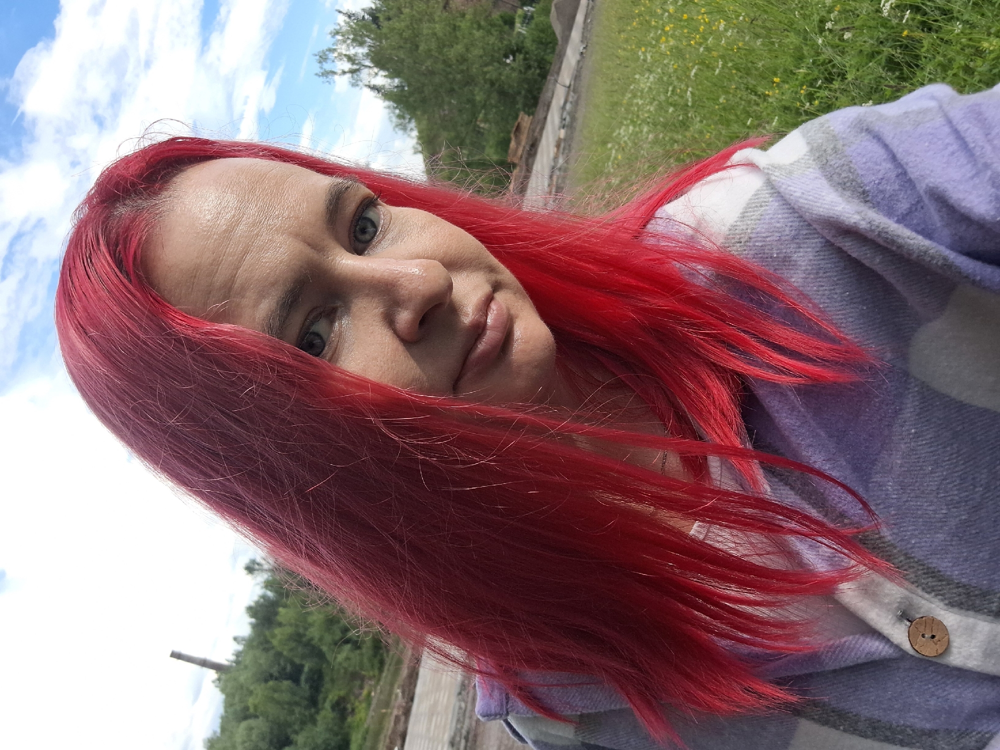 A medium shot selfie taken outdoors shows a woman with bright red hair, facing slightly to her right. She has light-colored eyes and her mouth is slightly open. She is wearing a light-colored shirt layered under a plaid jacket with stripes of purple, gray, and white. A wooden button is visible on the jacket. The background is a grassy field with some small white and yellow flowers. In the distance, there are trees, a cloudy sky, and a tall, thin industrial structure or smokestack. The scene appears to be located on the outskirts of Sim, Russia, possibly in an industrial or construction area with surrounding natural elements.