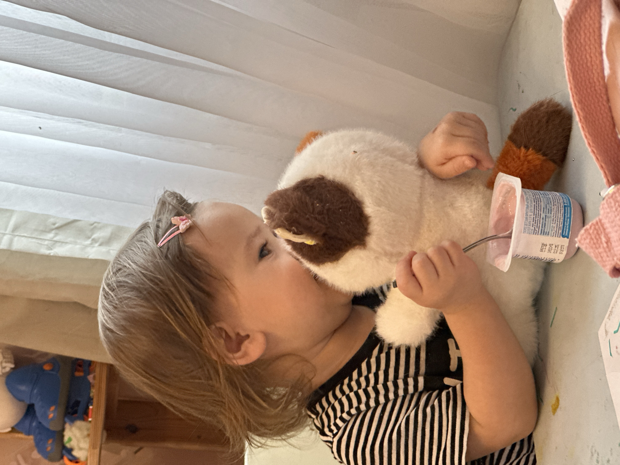 A toddler with blonde hair and a pink hair clip, wearing a black and white striped shirt, is holding and kissing a stuffed animal cat. The cat is white with a brown face and an orange and brown striped tail. The child is also holding a small container of yogurt with a spoon. The location is indoors, possibly a child's room, with a white window shade visible in the background. In the lower-left corner of the image are various colorful toys. There is no indication that the photo was taken in Kharnavy, Russia.