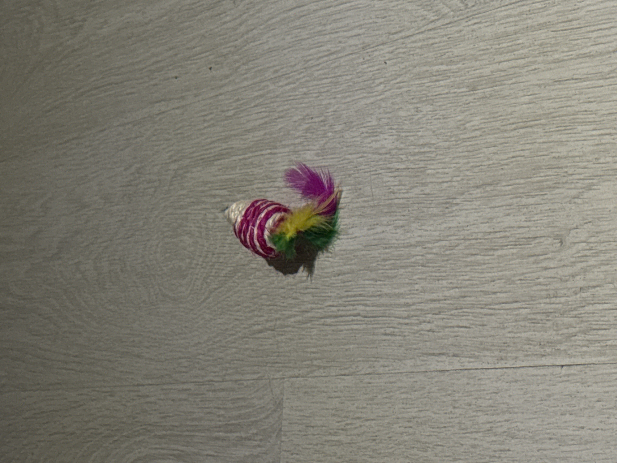 A toy mouse with pink and white stripes and a colorful feather tail lies on a light-colored wooden floor. The toy is approximately 2 inches long and is positioned diagonally on the floor, with its tail pointing towards the right. The floorboards run horizontally across the image. The image is well-lit, with a shadow cast by the toy to its right. The scene appears to be an indoor setting, likely a home. There are no people or other objects visible in the frame. There is no information about the location in Podkletnoe, Russia, as this is a generic toy and floor setting.