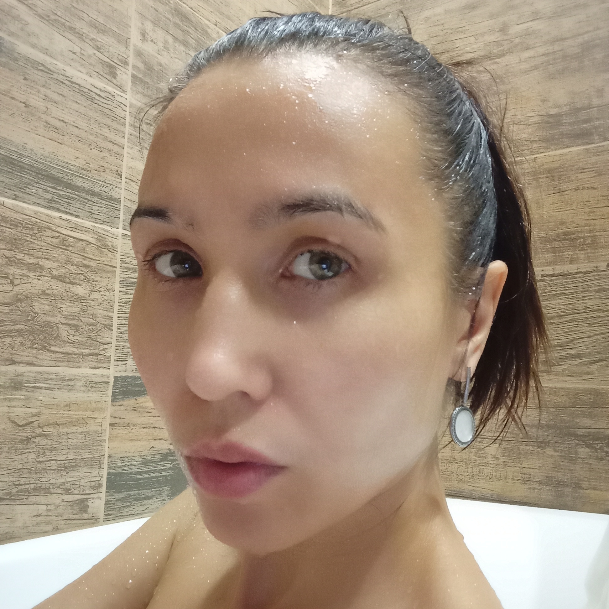 A woman with wet hair is in a bathtub. The bathroom has wood-patterned tile walls. The woman's hair is pulled back into a ponytail. She has fair skin and is wearing earrings. There is water visible on her face and upper body. There is no indication of the location of the city of Pervomaysky, Russia.