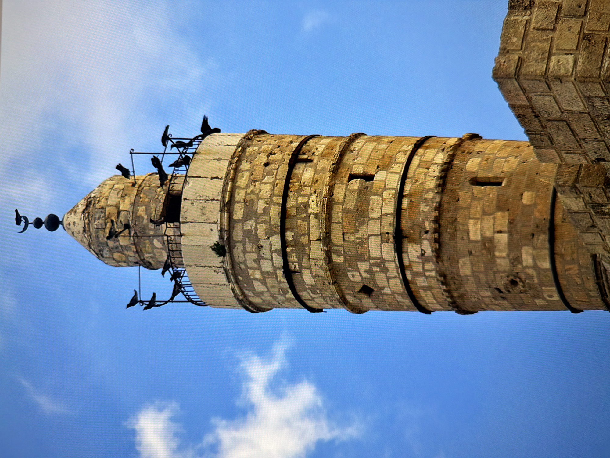 The image shows a stone tower with a conical roof against a bright blue sky with scattered clouds. Several birds are perched on a railing around the top of the tower and on the roof. The tower is constructed of large, weathered stone blocks and appears to be ancient. There is a crescent moon finial on top of the roof. In the foreground, to the right, are more stone blocks, suggesting the tower is part of a larger structure or wall. The lighting suggests it is daytime. The image does not provide any information about a specific location in Kyiv, Ukraine.