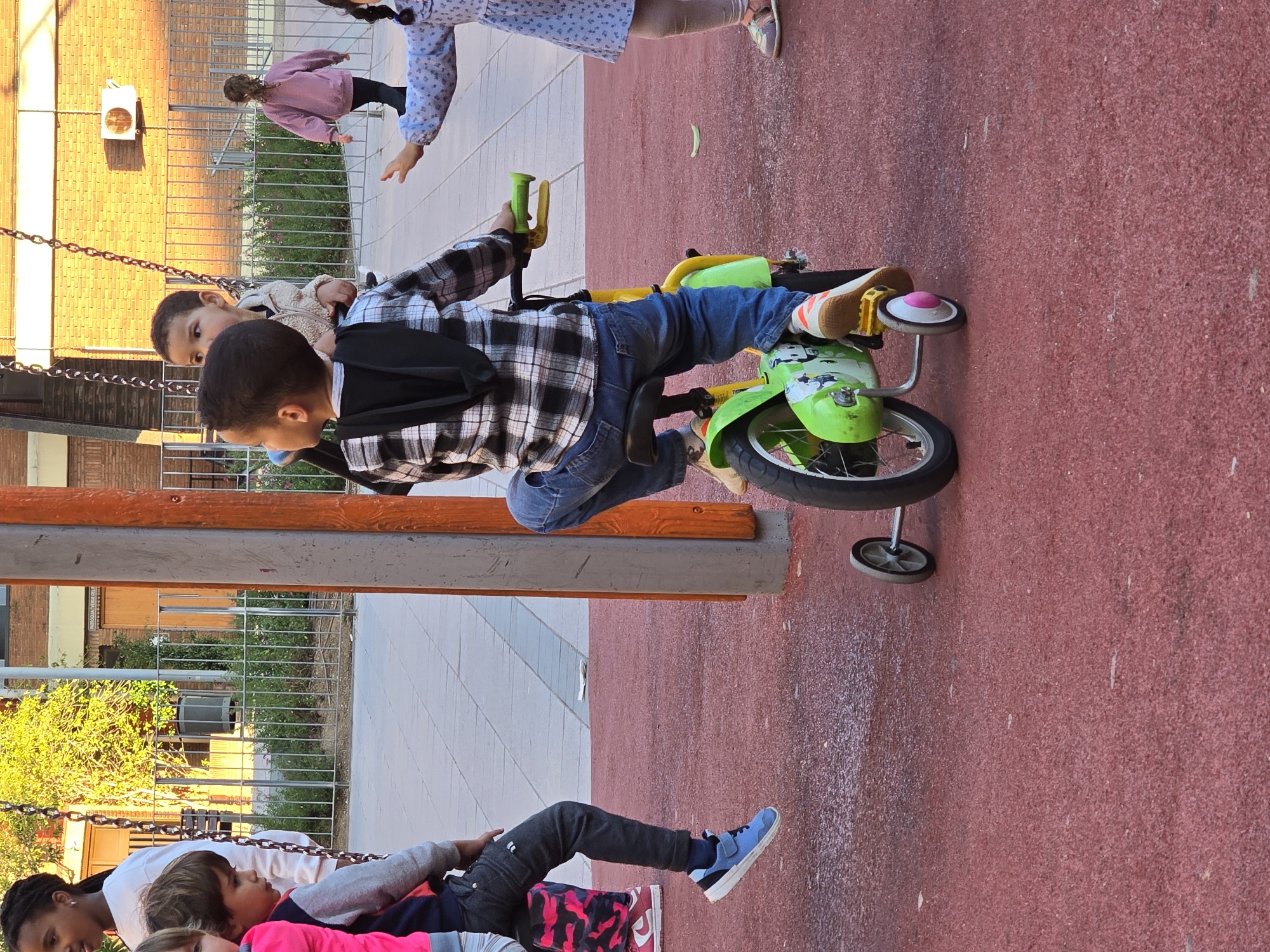 A group of children interacts on a playground in Cornellà De Llobregat, Spain. The ground is a red, textured surface. One child sits on a green and yellow bicycle with training wheels. The child wears a plaid shirt, blue jeans, and a backpack. Another child stands nearby, and two more are on swings. One child in a pink shirt is lying down on the swing, with two other children lying with them. A building with a brick facade and an air conditioning unit is in the background. A metal fence and greenery are visible behind the swings.