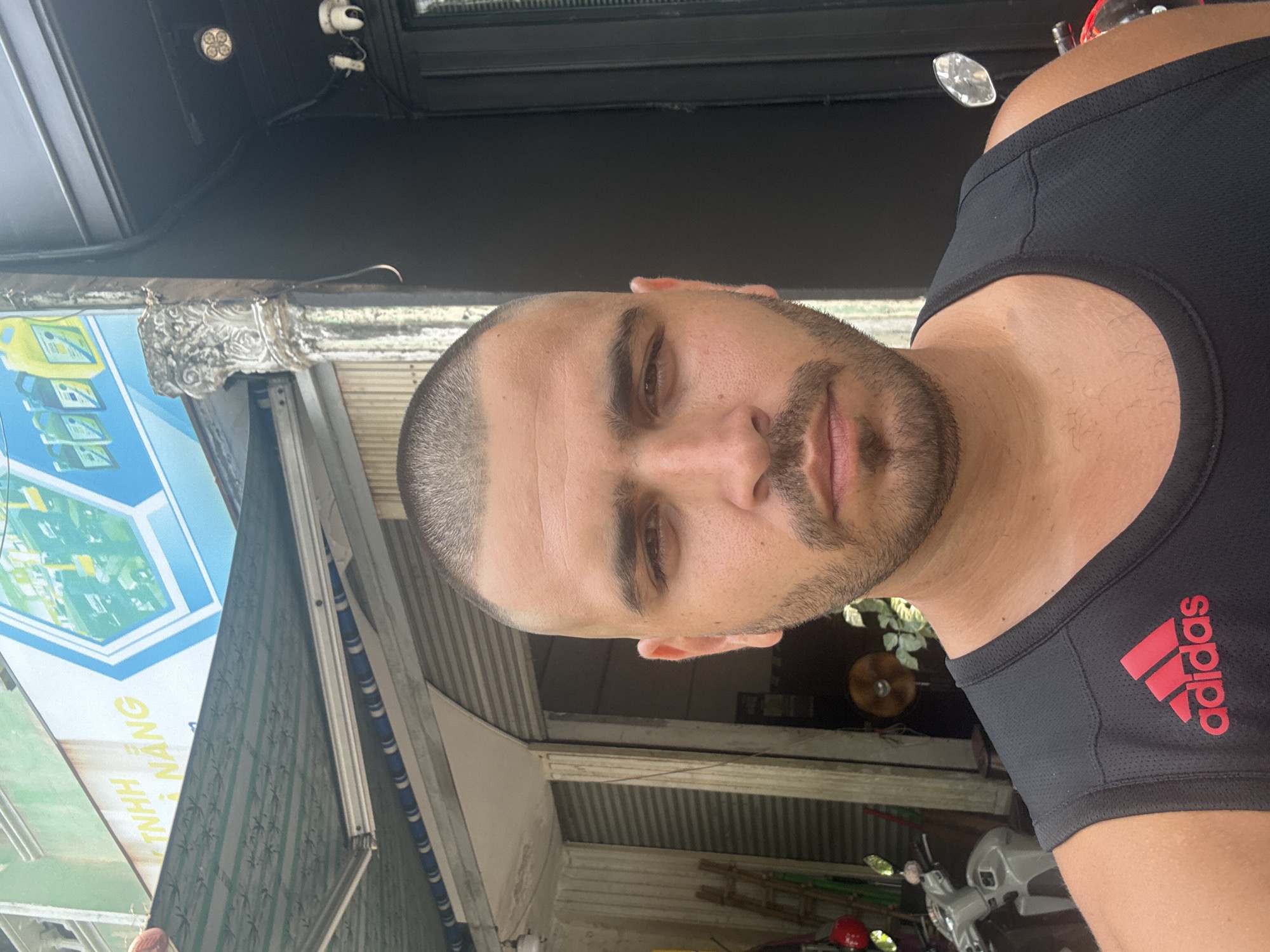 A medium close-up angled self-portrait shows a man with a shaved head, brown eyes, and a trimmed beard wearing a black Adidas tank top. The background suggests an outdoor setting. An awning with blue and white panels displays the words "TNHH NANG". Behind the man, corrugated metal and a scooter are visible, typical of urban scenes in Vietnam. Given the signage and architectural details, the location is likely Đà Nẵng, Vietnam. The lighting is ambient daylight. There is no action visible.