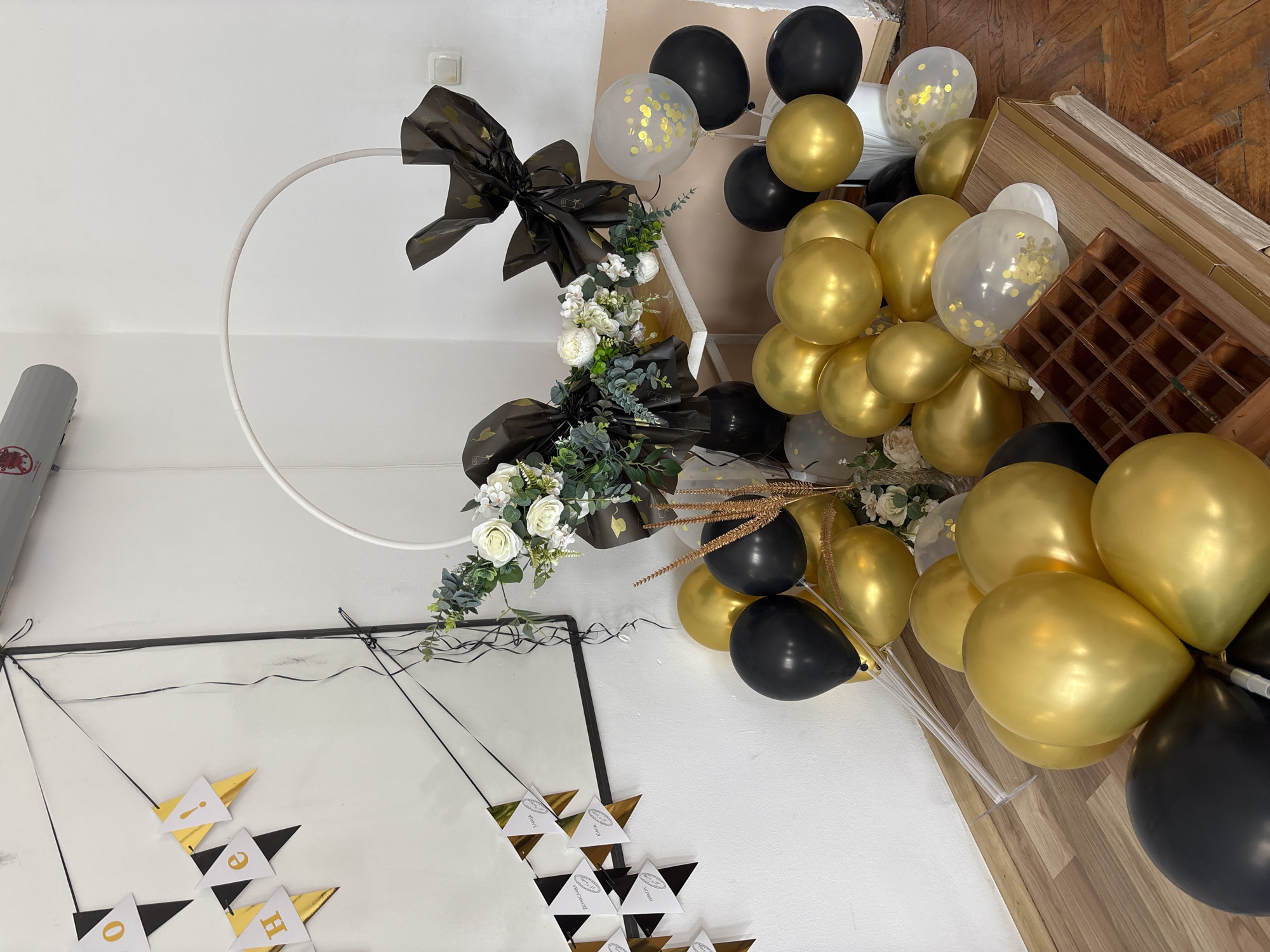 The image shows an indoor decorative setup. There's a white circular frame adorned with black bows, white roses, and greenery. Adjacent to the frame is a cluster of gold, black, and clear balloons with gold confetti inside some. A "WELCOME" banner is visible in the lower left corner. The scene appears to be set against a wall with what looks like a mirror and a wooden background with an empty tray-like container on the right. The location is not specified and cannot be determined based solely on the image content. No people or actions are visible.