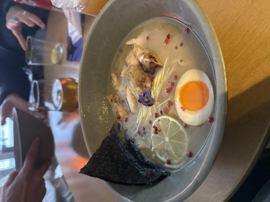 The image depicts an indoor dining scene at Ramen Beograd (Ramen to je ljubav) located at Dobračina 59b, Dorćol, Belgrade, Serbia. The primary subject is a large, light grey ceramic bowl filled with ramen-style soup, positioned centrally in the foreground on a warm-toned wooden table.
The ramen bowl contains a light-colored broth, noodles, several irregular pieces of seared or grilled meat (likely chicken), and thinly sliced white ginger or daikon. A dark, triangular piece of nori seaweed stands upright along the left rim. Garnishes include a round slice of green lime, numerous small red peppercorns (identified as pink peppercorns) scattered across the surface, and a small cluster of purple edible flowers positioned near the center. A soft-boiled egg, sliced longitudinally to expose a vibrant orange yolk and white albumen, is situated in the lower right portion of the bowl.
The background, partially blurred, reveals additional elements on the wooden table and parts of other diners. On the left, hands are holding a smaller, light-colored bowl. To the right of the ramen bowl, a clear glass contains an amber liquid, consistent with beer. Further to the right, an empty clear drinking glass, a red and white Coca-Cola can, and a glass with an orange-hued beverage are visible. The scene is illuminated by ambient light, suggesting an indoor restaurant setting with external light sources contributing to reflections in the background.