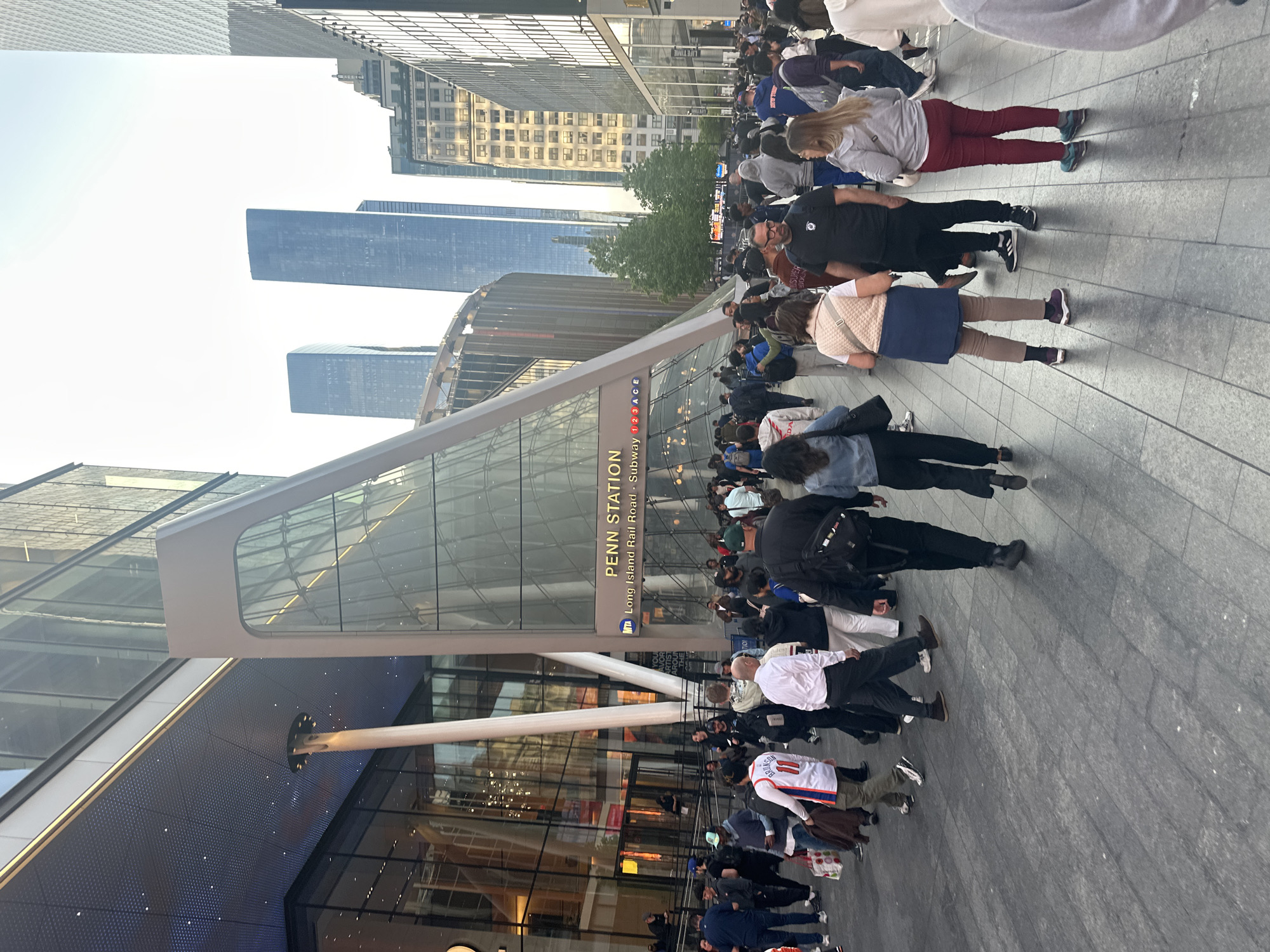 This is an outdoor urban scene depicting a bustling crowd of people in front of a modern glass and steel building. The structure features a distinctive angular, possibly triangular, glass entrance. Tall skyscrapers dominate the background, characteristic of a major city environment.
Numerous pedestrians, appearing to be commuters, are walking across the paved area in front of the building. They are dressed in various casual and semi-casual attire, moving in different directions, likely entering or exiting the building. The overall atmosphere suggests a busy transportation hub.
The lighting indicates late afternoon or early evening, with soft, natural light and visible shadows, under what appears to be a clear sky.
Prominently displayed on the building's facade is a sign with the text "PENN STATION", along with "Long Island Rail Road" and "Subway 1 2 3 A C E". An "MTA" logo is also visible, confirming the location as Penn Station in New York City, United States of America, likely depicting the Moynihan Train Hall entrance. People appear focused on their movement, with no strong emotions discernible among the crowd.