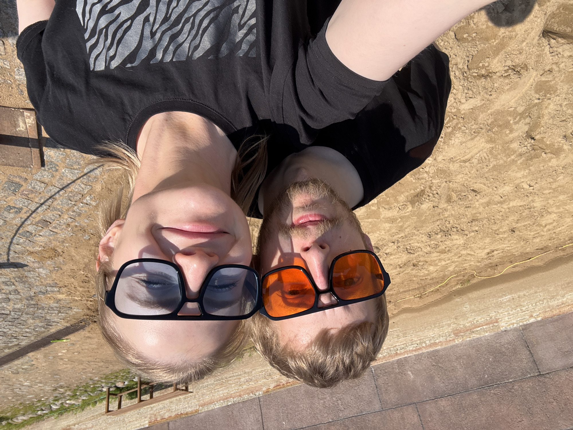 The image shows a man and a woman lying on their backs next to each other, both wearing sunglasses. The woman is on the left wearing dark-framed sunglasses with blue tint, while the man is on the right with orange-tinted sunglasses. They are lying close together, facing the camera. They appear to be lying on a brick pavement adjacent to a sandy area, suggesting a beach or an urban recreation area with sand. They both wear dark, casual tops. Based on the visual cues like architecture style, the setting appears to be the beach in Saint Petersburg, Russia.