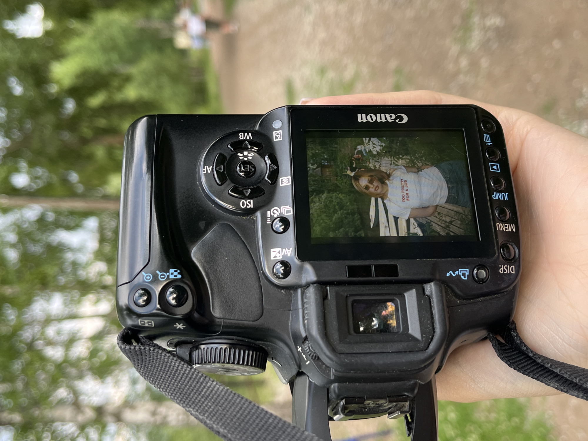 The image shows the back of a black Canon DSLR camera being held by a person's hand. The camera's LCD screen displays a photograph of a young woman wearing a white t-shirt with red text that says "TOO PRETTY FOR A JOB," denim shorts, and sunglasses. She is standing outdoors with some greenery and boats in the background. The camera itself has various buttons and a dial visible. The background of the main image is a blurred outdoor scene with trees and a dirt path. Two blurred figures are walking away in the distance on the path. The location is identified as Chaikovsky, Russia, based on context not present in the image itself. The scene appears to be a casual outdoor setting, possibly a park or recreational area.