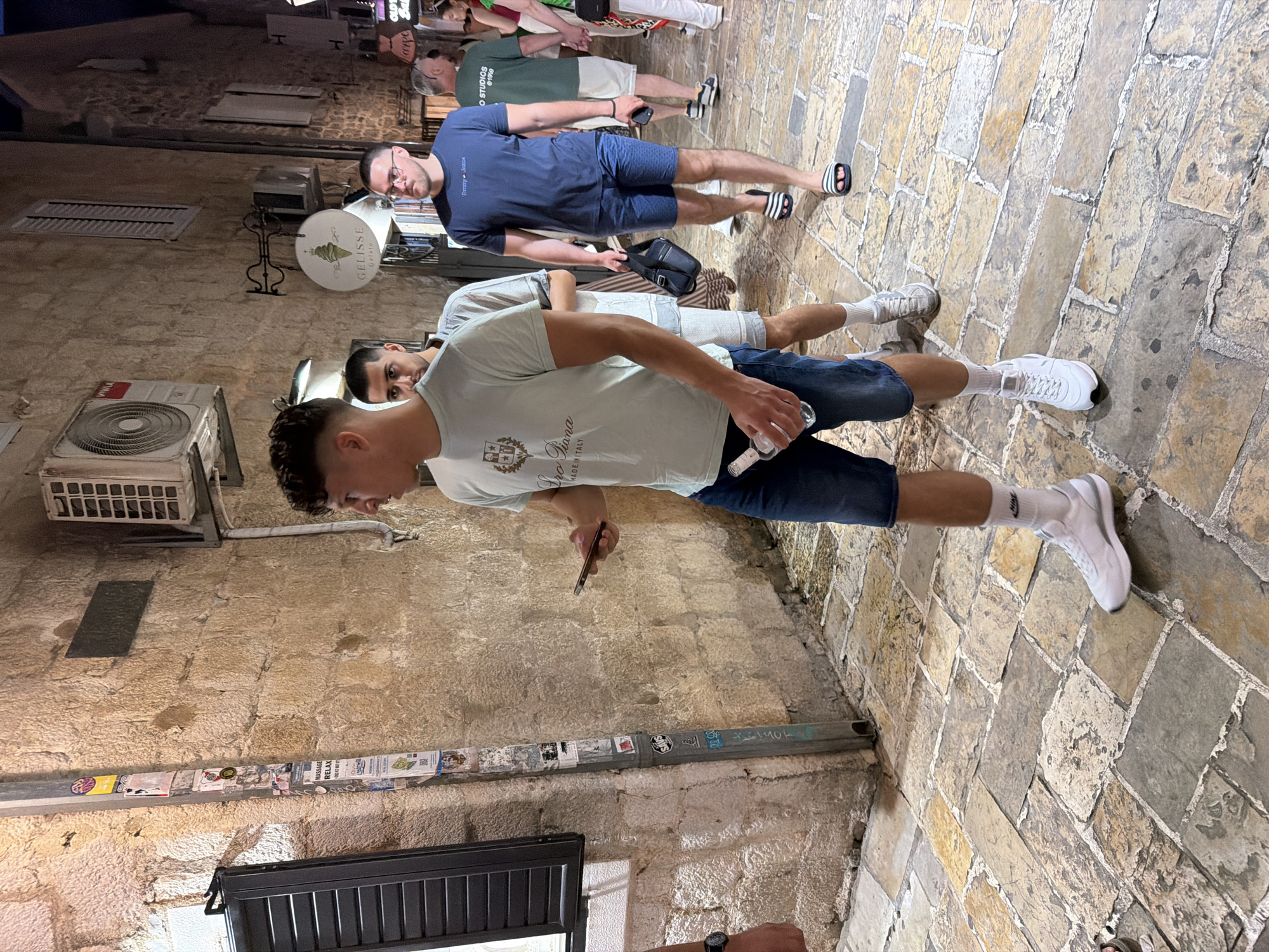 In the city of Budva, Montenegro, a group of young men walks along a cobblestone street at night. The street is lined with buildings constructed of rough stone, illuminated by the warm glow of streetlights and interior lights from shops. In the foreground, a man wearing a light blue t-shirt and denim shorts is holding a smartphone in his left hand and a plastic bottle in his right. He is walking alongside another man in a grey t-shirt and shorts. Behind them, a man in a blue t-shirt and dark shorts carries a black crossbody bag and wears flip-flops. Further down the street, other individuals are visible, including a man in a green t-shirt and a woman in a white dress with a red scarf. Signs for "Gelisse Gelato" and "Tommy Jeans" are visible on the buildings, indicating commercial establishments. An air conditioning unit is mounted on the stone wall of one building. The overall atmosphere suggests a lively evening in a European coastal town.
