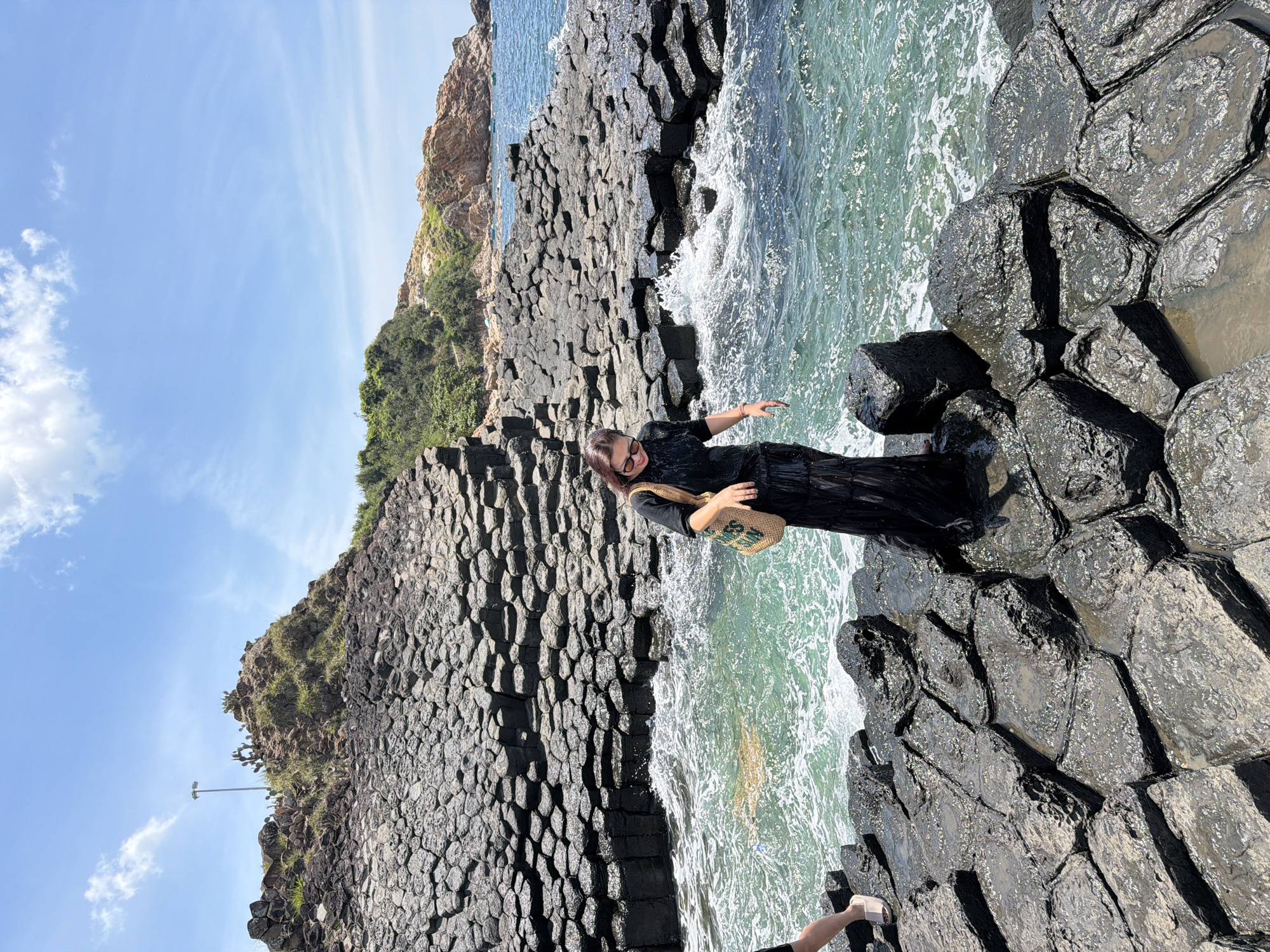 A woman stands on a rocky shoreline near Phú Lương, Vietnam. The rocks are hexagonal basalt columns, a geological formation. She is wearing a long black dress and sunglasses. She carries a woven bag over her shoulder. The ocean water is turquoise and wavy, with some splashing onto the rocks. In the background, there are more hexagonal rock formations leading up to a vegetated hillside. The sky is blue with a few white clouds.