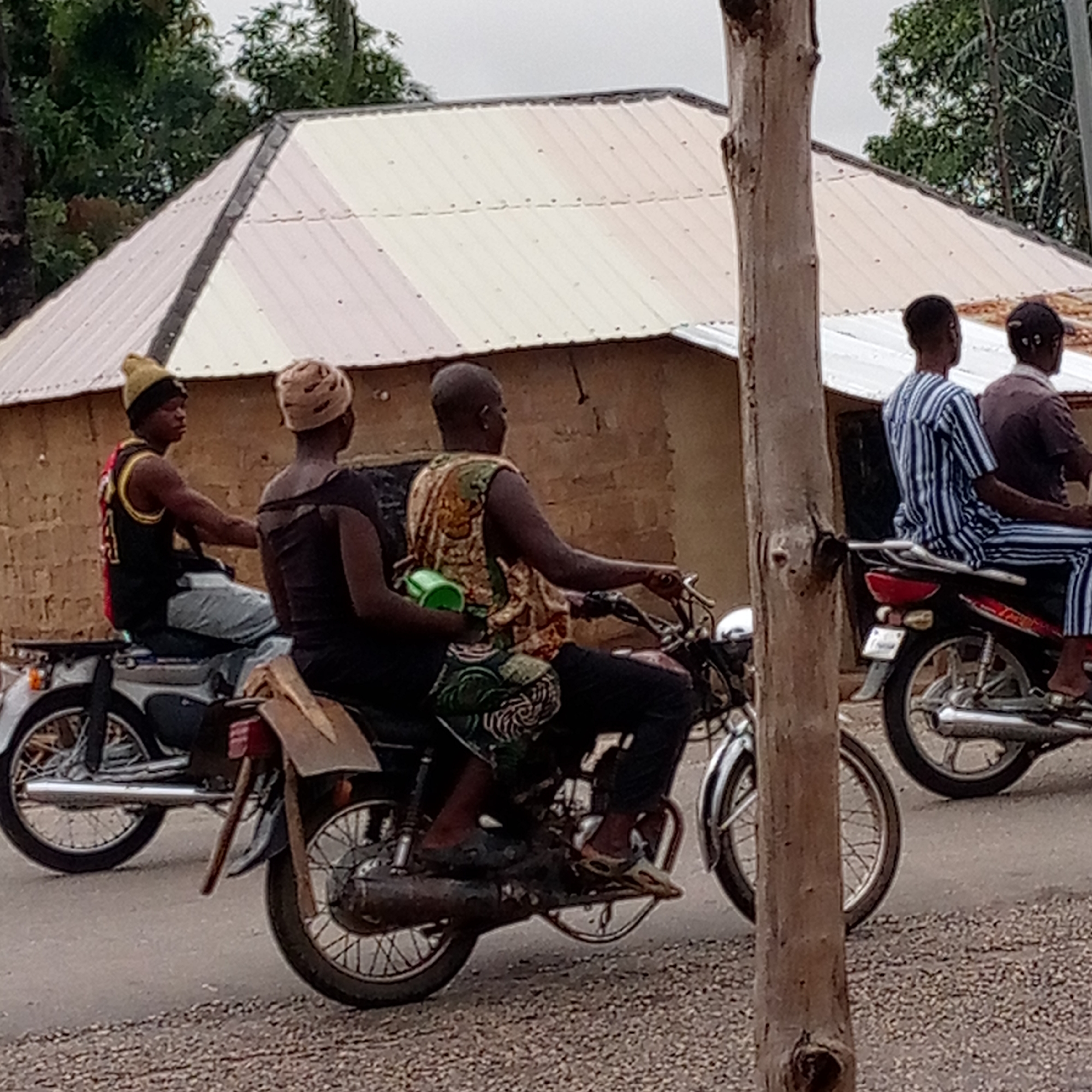 The scene is in Mararaba, Nigeria. Three motorcycles are on a road, partially obscured by a tree trunk in the foreground. The first motorcycle has one rider wearing a black and red tank top and a yellow knit cap. The second motorcycle has two riders, the driver wearing a patterned tank top and a passenger holding a green cup. A shovel is strapped to the back of the second motorcycle. The third motorcycle has two riders, both wearing striped clothing. Behind the motorcycles is a building with light-colored corrugated metal roofing. The sky is overcast.