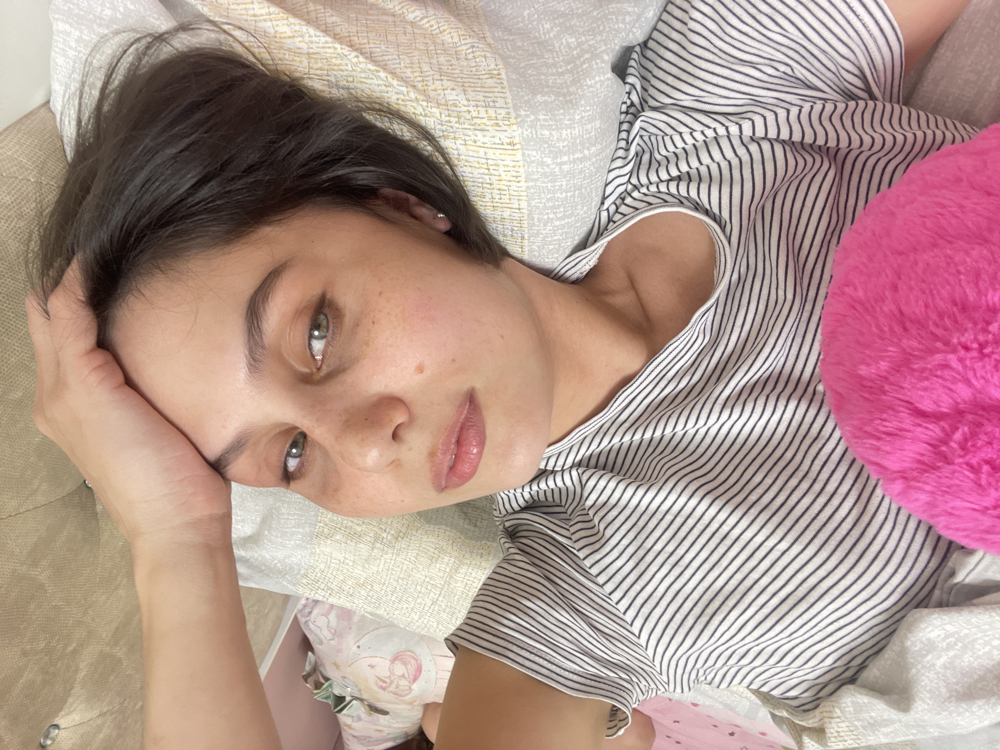 A young woman lies on a bed. Her head rests on a pillow, and her right hand is positioned near her forehead. She is wearing a striped shirt. A pink, fluffy object is adjacent to her. The background includes bedding and hints of furniture. The location is unknown and cannot be determined from the image.