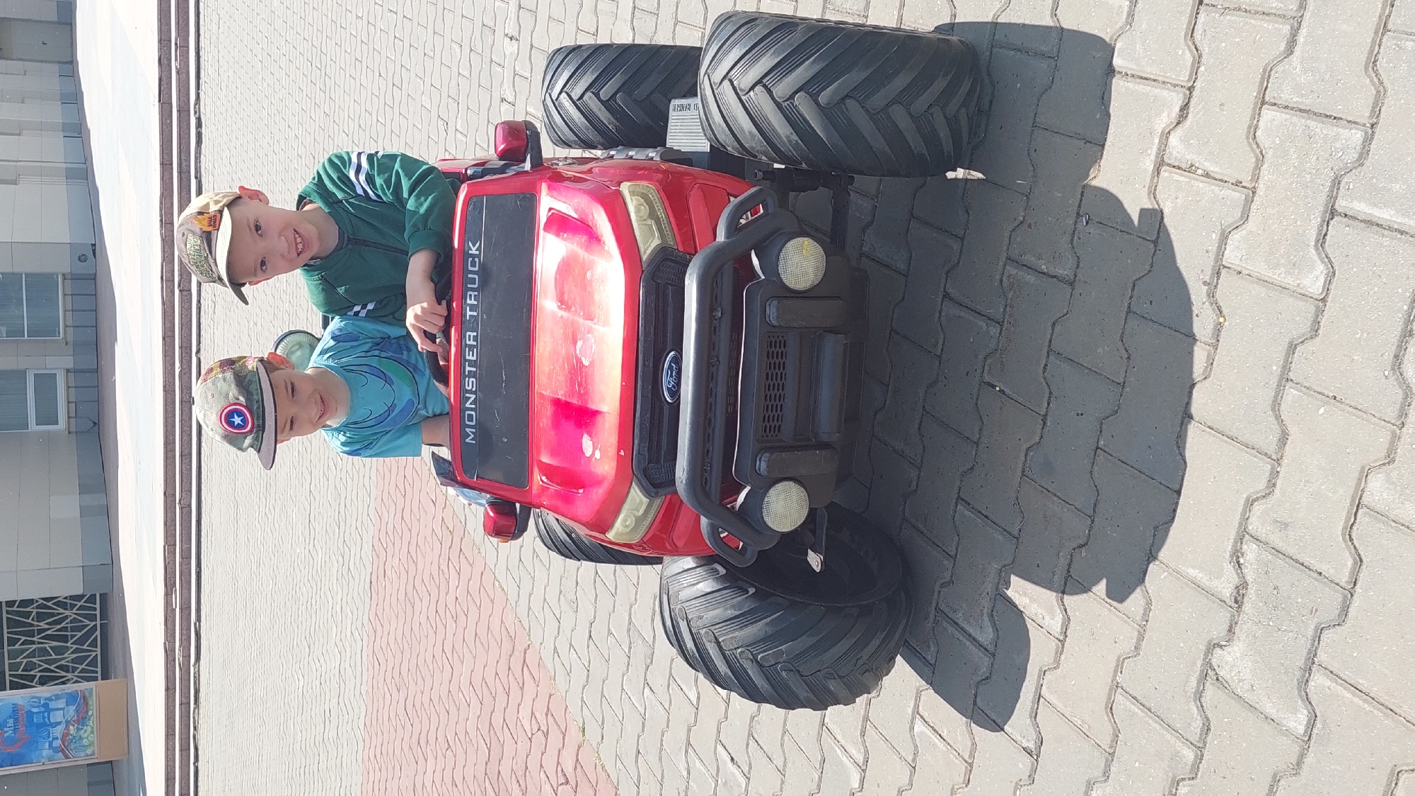 Two young boys, wearing baseball caps, are seated in a red, ride-on toy monster truck. The boy on the left is wearing a blue t-shirt with a cartoon character on it. The boy on the right is wearing a green jacket with white stripes on the sleeves. They are smiling and looking towards the camera. The toy truck is parked on a paved surface made of interlocking bricks, likely in an outdoor public space, such as a park or a square in Komsomolsk-on-Amur, Russia, as indicated by contextual information. The background shows a building with windows and a poster on the left side. The sun is casting shadows, suggesting it is daytime.