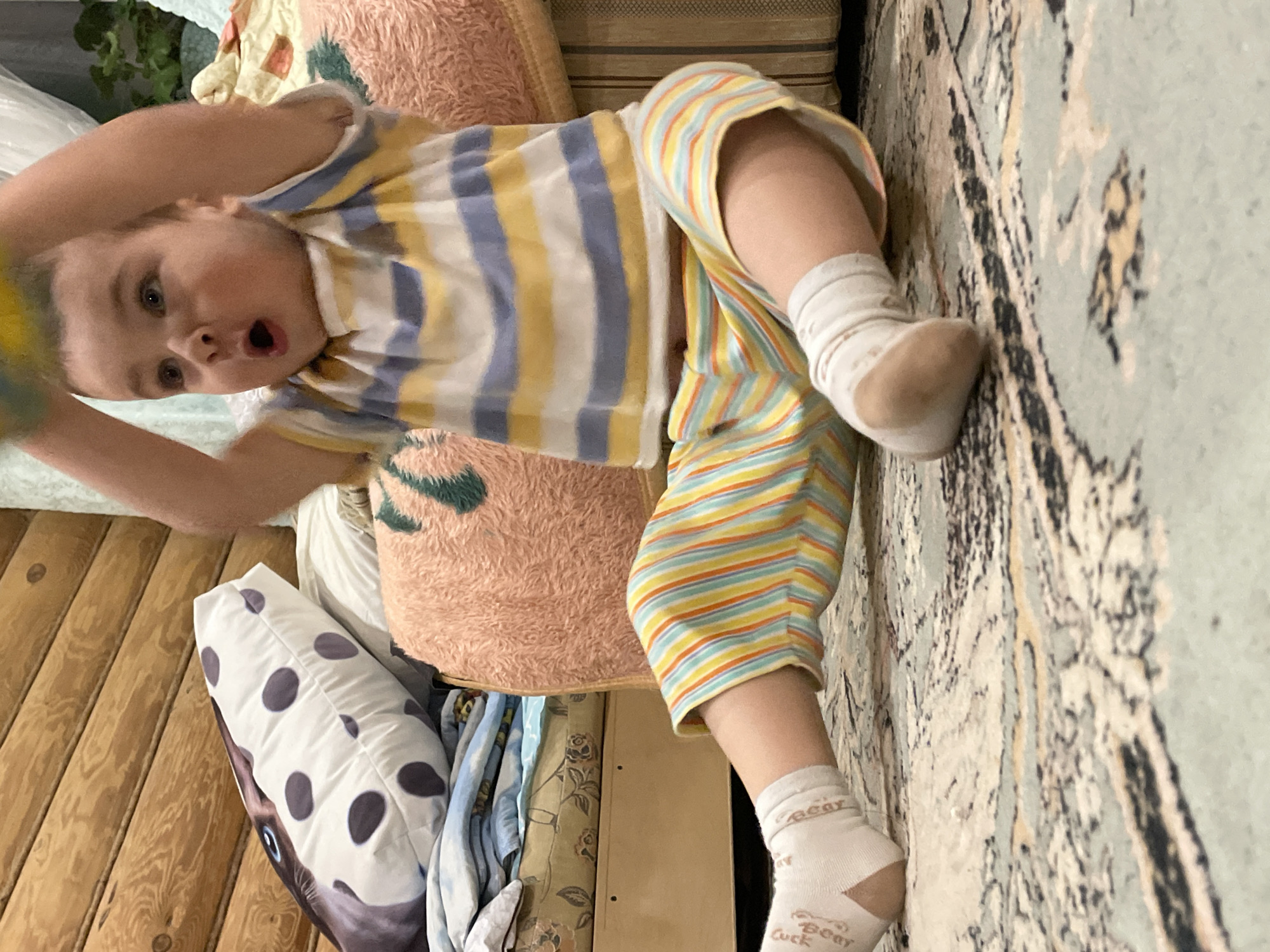 A child lies on a carpet, propped up by pillows and possibly furniture. The child is wearing a striped shirt and pants, and white socks with text visible on the ankle. The environment includes wooden paneling on one wall, and the carpet has a complex pattern. Other cushions or bedding are present. The location is not discernible based on the visual information provided.