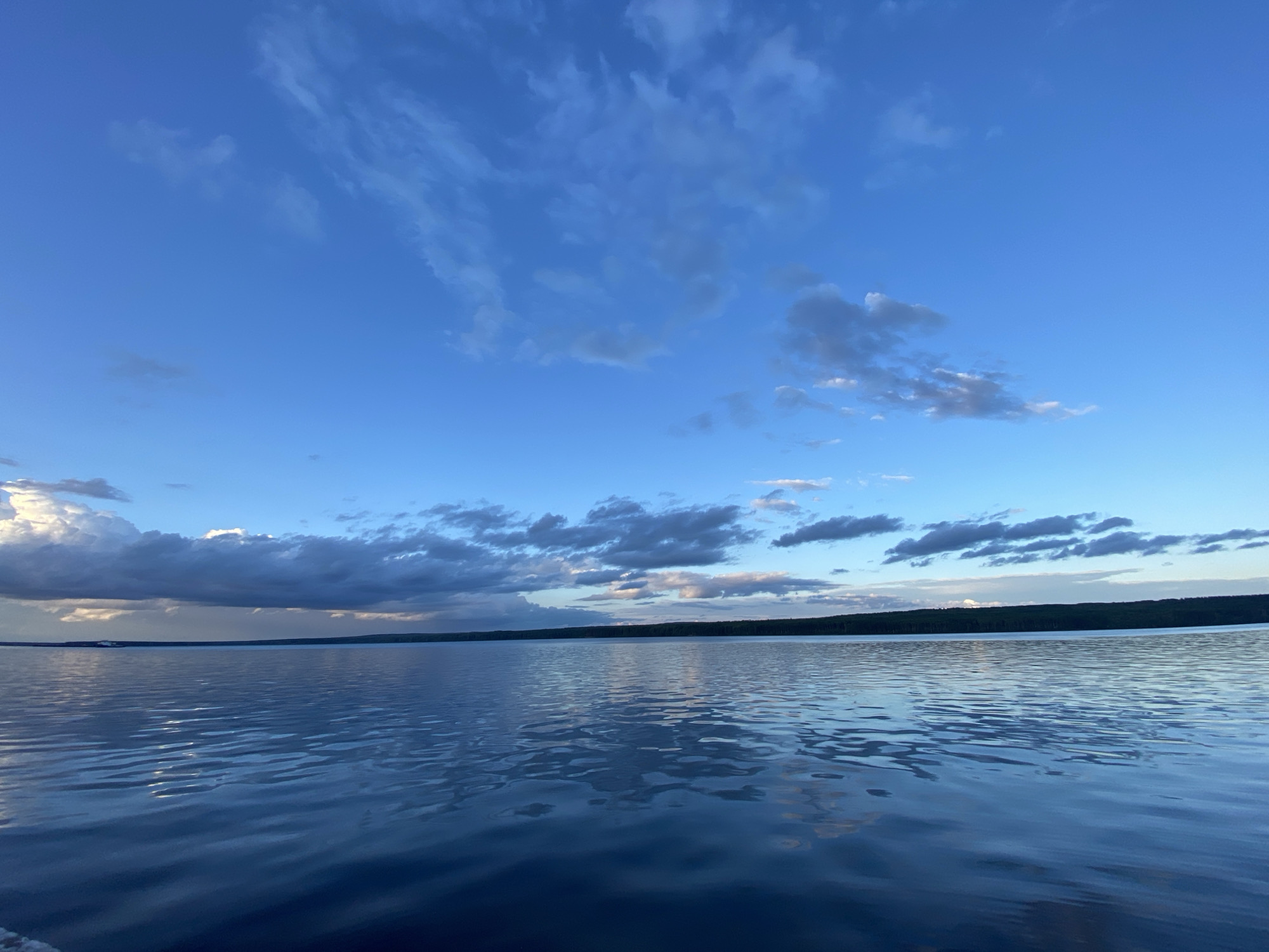 The image shows a wide, calm body of water, likely a lake or reservoir, with gentle ripples reflecting the sky. The sky is a vibrant blue with scattered white and gray clouds. The horizon line is low, separating the water from a dark, tree-lined shoreline. The overall impression is one of tranquility and natural beauty. This scene could be characteristic of the Kama River near the city of Chaikovsky, Russia, which is known for its expansive water bodies and scenic landscapes. There are no people or man-made structures immediately visible, suggesting a natural, undisturbed environment.