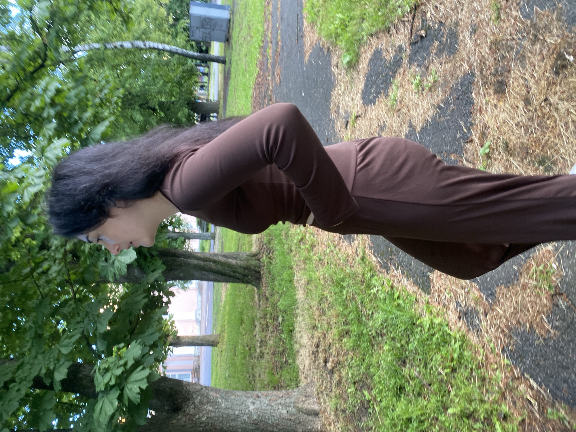 A woman stands on a grassy area next to a paved path. The woman has long, dark hair, fair skin and is wearing a brown, long-sleeved jumpsuit. She is facing left, with her head tilted downward slightly. In the background, there are trees, grass, and a building, suggesting a park-like setting. The location is likely Poltava, Ukraine, as that was specified as the location. The surface is a mix of grass and patches of dry leaves.