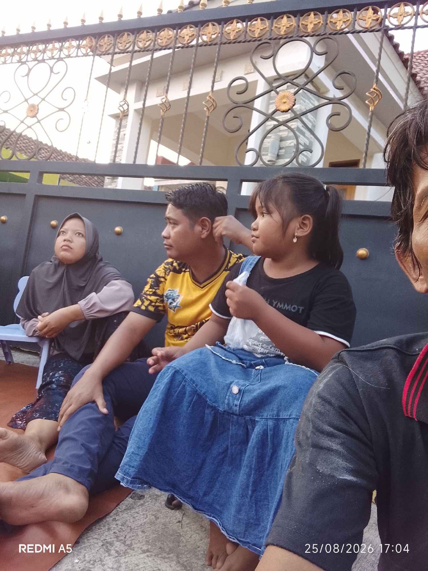 The image depicts a group of four individuals, including two adults and two children, seated outdoors in what appears to be a residential area in Semanding, Indonesia. The individuals are positioned in front of a decorative black metal gate with gold embellishments, which separates them from a building with a tiled roof and white walls.
On the left, a woman wearing a dark grey hijab and a light grey long-sleeved shirt is seated on a light blue chair. Her hands are clasped in her lap, and she is looking upwards with a thoughtful expression. Next to her, a man in a yellow and black patterned t-shirt is seated, looking to his right. A young girl, wearing a black t-shirt with "LYMMOT" printed on it and a blue denim skirt, is seated to the man's right, holding a white plastic bag. Her hair is pulled back in a ponytail, and she is wearing a small hoop earring. To the far right of the frame, partially visible, is another individual wearing a black t-shirt with red and black striped detailing around the neckline.
The ground beneath them is a mix of reddish-brown earth and grey concrete. The overall lighting suggests it is daytime, possibly late afternoon given the timestamp "25/08/2026 17:04" visible in the bottom right corner. The presence of a gate and building indicates a domestic setting. The individuals appear to be interacting casually, suggesting a family gathering or social occasion. The atmospheric conditions are not discernable from the image.