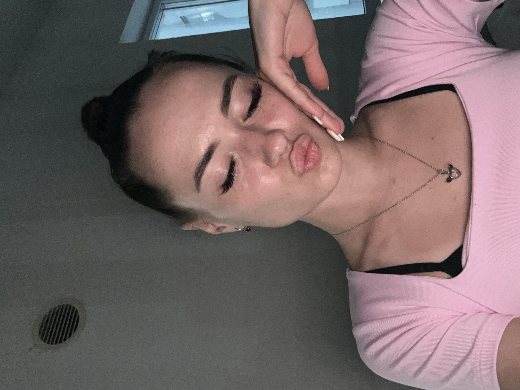 An overhead, medium shot captures a young woman in a dimly lit room, her head tilted and eyes closed in a puckered kiss pose. She rests her cheek on her hand, her fingers splayed. She wears a pink top over a black bra and a silver necklace with a heart-shaped pendant. Her hair is pulled into a bun. In the background, a circular vent is visible on the wall. A window with blue light can be seen on the right. The scene appears to be indoors.