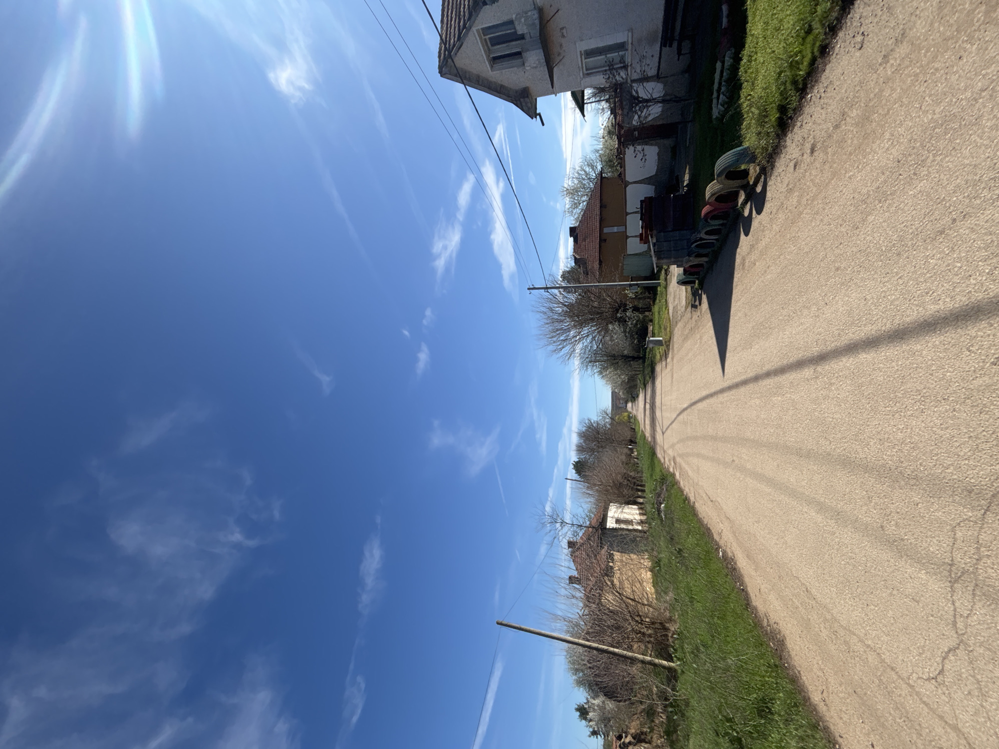 The image depicts a rural street scene in Lehchevo, Bulgaria. The road is unpaved, with grass and some bare trees lining the right side. Houses are visible on both sides of the street, some appearing old and in need of repair. A stack of used tires is visible along the roadside near a house. A clear blue sky with wispy clouds occupies the upper portion of the image. The perspective suggests the photo was taken at ground level, with the camera tilted slightly. No people are visible in the frame. Power lines run overhead.