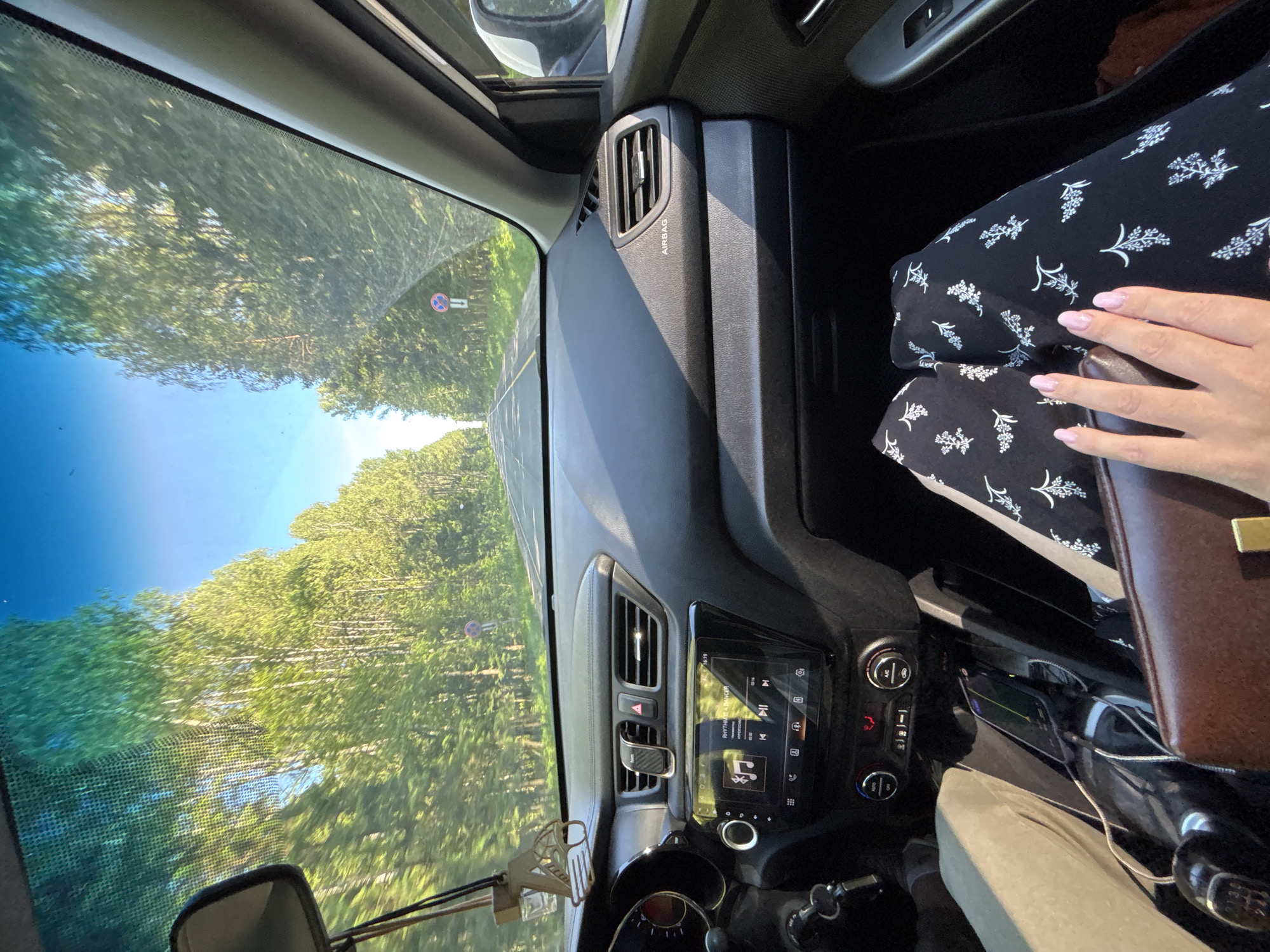 The image captures the interior of a vehicle from a front passenger perspective, looking forward through the windshield onto a tree-lined road in Rusanovo, Russia, under clear blue skies.
The vehicle's dashboard is black, with a central infotainment system displaying "RHYTHME A DANCER" by "SNAP!" playing at 02:32 of 03:16, with the current time noted as 16:19. A phone mount is affixed to the central air vents, and hazard lights are visible. On the passenger side, the dashboard is labeled "AIRBAG" above an air vent.
The driver's left lower leg and foot, clad in light-colored trousers, are visible on the left. A manual gear shift with a black knob is also visible to the lower left. In the center console area, a mobile phone is charging via a cable.
A passenger's legs, wearing dark pants with a white floral pattern, are visible on the right. The passenger's right hand, with light pink nail polish, rests on a brown leather-like purse with a metallic clasp.
Through the windshield, a two-lane asphalt road with a dashed yellow center line and a solid white shoulder line recedes into the distance, bordered by dense rows of tall, slender deciduous trees with vibrant green foliage. A circular road sign with a red border, blue background, and a red 'X' over a white 'H' (indicating "No Stopping" or "No Parking") is visible on the right side of the road further down. A car air freshener hangs from the rearview mirror post in the upper left.