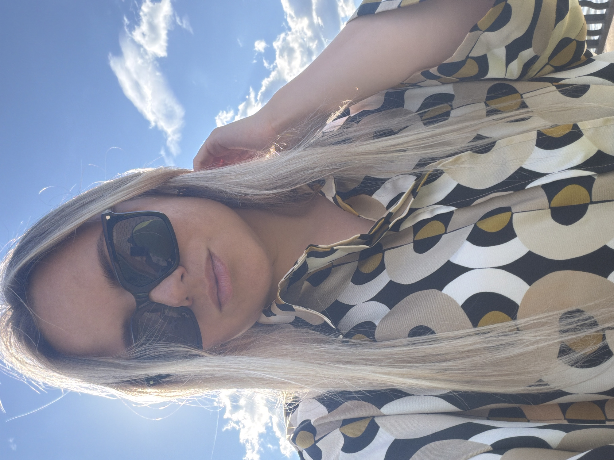 A single young adult female with long, straight, blonde hair is depicted in a close-up, outdoors selfie. She is wearing black, rectangular sunglasses, which reflect bright light and elements of the sky. Her lips are slightly parted. She is dressed in a collared shirt featuring a geometric pattern composed of concentric and overlapping circles in shades of black, white, beige, and mustard yellow. Her right hand is visible on the right side of the frame, touching her hair near her ear. The background consists of a clear blue sky with scattered white cumulus clouds. Bright sunlight emanates from behind and to the left of the subject, creating a strong backlight and highlights on her hair. The scene is set in Votkinsk, Russia.