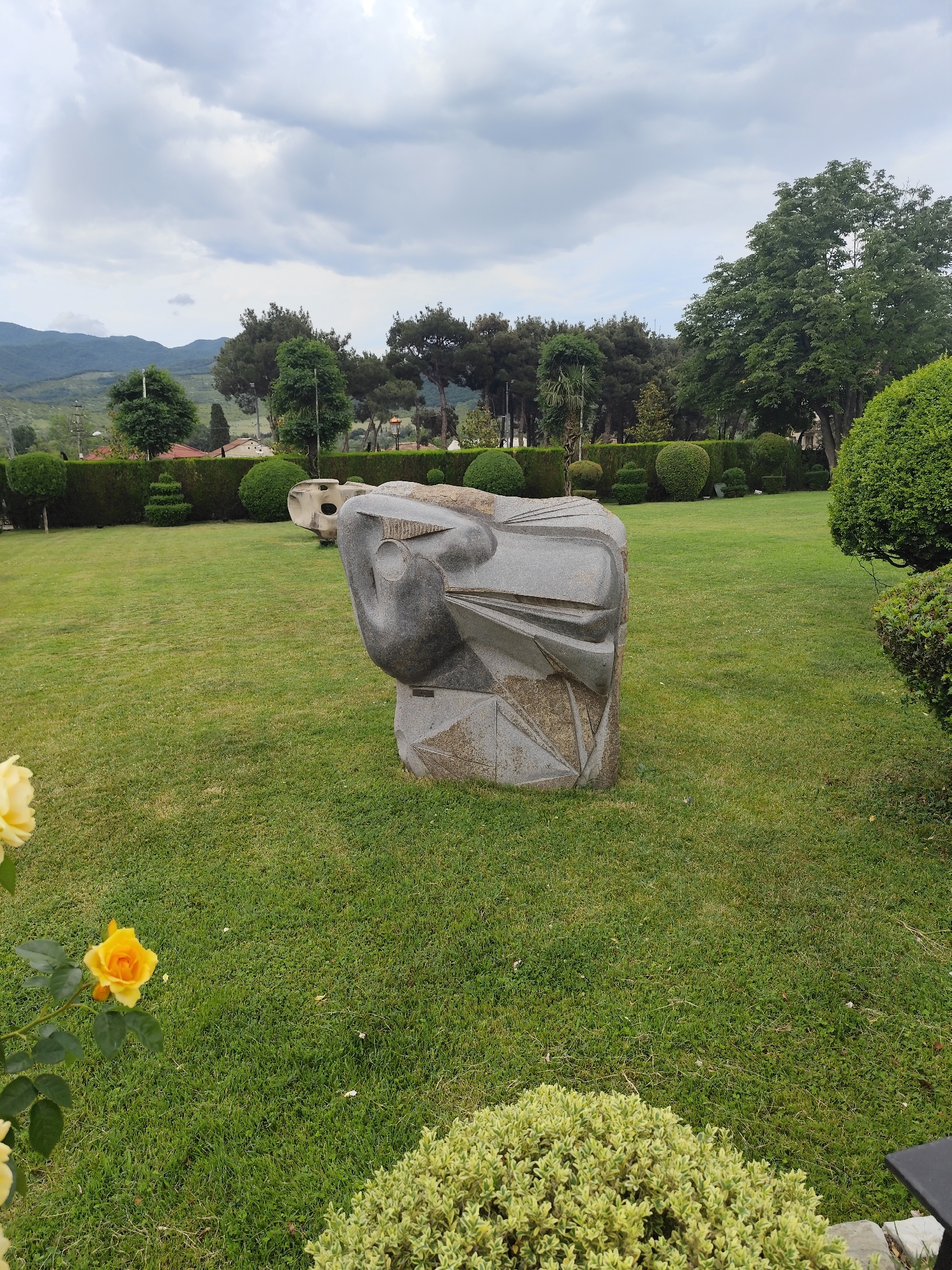 A wide shot captures a stone sculpture in a verdant park in Mtskheta, Georgia. The sculpture, composed of angular, abstract stone elements, stands prominently on a manicured green lawn. In the foreground, a yellow rose bush with two blooming yellow roses adds a touch of color to the left side of the frame. The background features a neatly trimmed hedge, various trees, and in the distance, rolling hills under a cloudy sky. Another abstract stone element is visible further back in the park. The scene depicts an outdoor public space, likely a garden or park, intended for aesthetic appreciation.