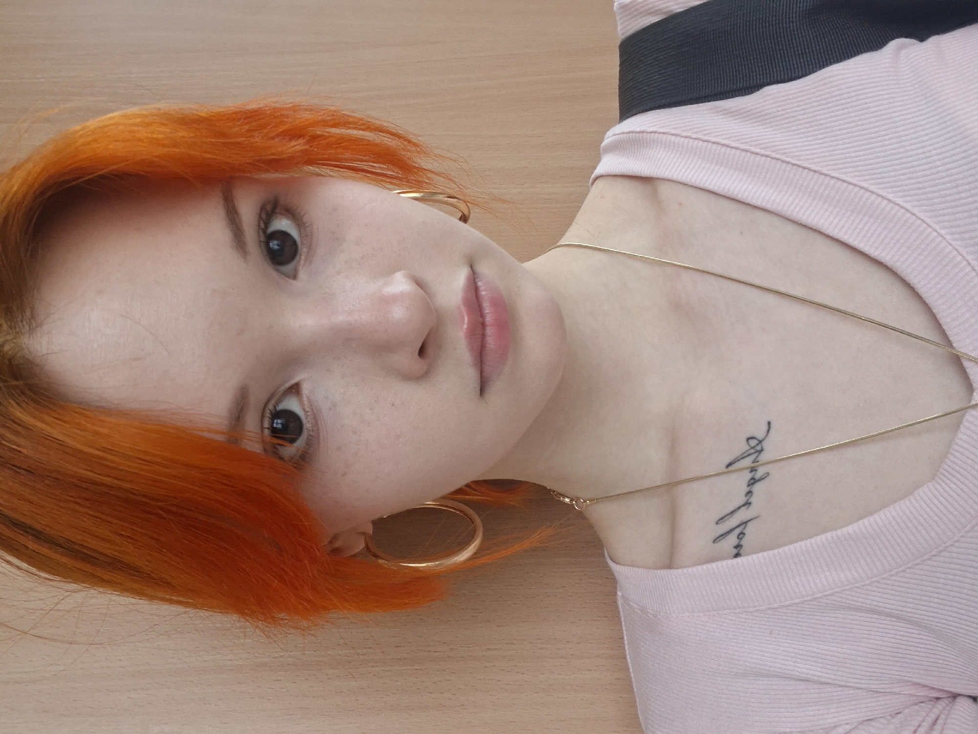 A fair-skinned person with freckles and vibrant orange, short hair is facing the camera, tilted slightly to their right. They have dark eyes, full lips, and are wearing large gold hoop earrings. A thin gold necklace adorns their neck. They are wearing a light pink, textured, crew-neck shirt. A dark strap, possibly from a bag, is visible over their shoulder. A tattoo is present on their upper chest near the collarbone. The background is a light brown surface, possibly a wooden table or desk.
There is no information available to determine that this picture was taken in Chaikovsky, Russia.