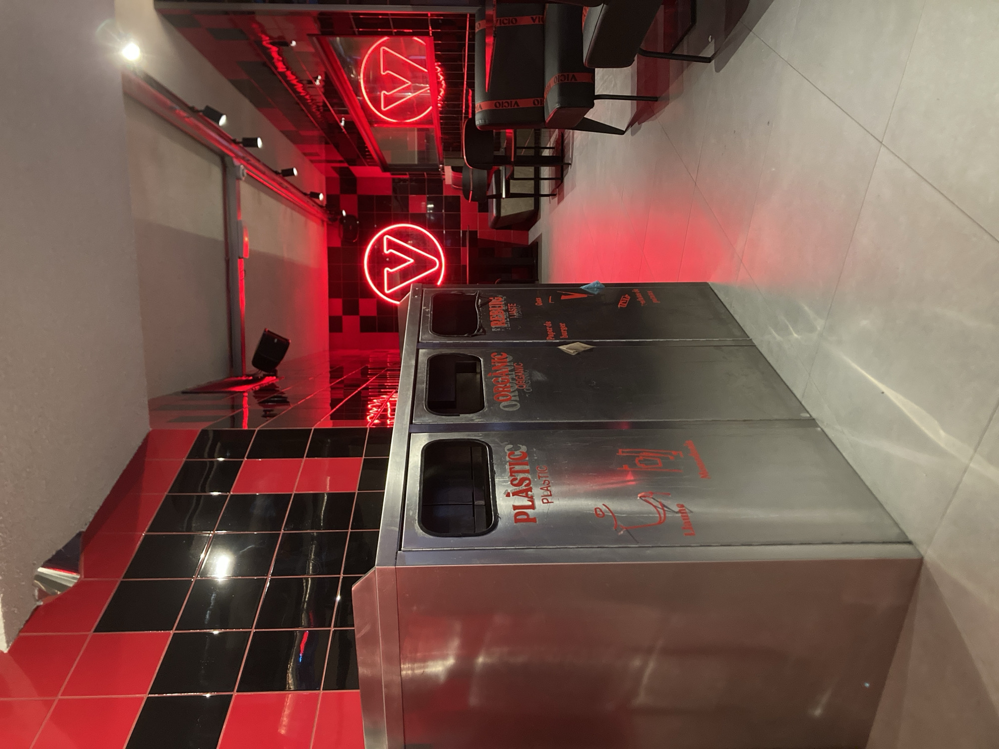 The image depicts the interior of a Vicio burger restaurant in Barcelona, Spain. The restaurant features a bold, modern design with red and black checkered tiles on the walls and neon red lighting, including the prominent "V" logo of the establishment. In the foreground, a set of three stainless steel recycling bins is visible, labeled "PLASTICO," "ORGANIC," and "REJE." The bins have slots for waste disposal and feature red graphics and text, including recycling symbols and descriptions of acceptable waste. The floor is tiled in a light color, reflecting the red neon lights. In the background, dark seating with red accents, likely booths and chairs, is arranged around tables. The overall atmosphere is industrial and contemporary, amplified by the strong color scheme and lighting. There are no people visible in the image.