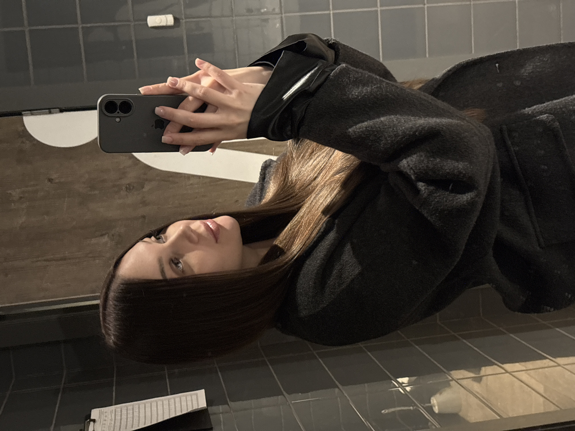 A woman with long brown hair is holding a gray smartphone in her hands, seemingly taking a selfie. She is wearing a dark gray coat with shiny black leather cuffs. The background consists of gray tiled walls and a white semi-circular object attached to the wall, possibly a sink or part of the bathroom fixture. There is also a white object visible near the top of the wall. A document is lying on the table. The location appears to be inside a restroom or bathroom in an unknown city.