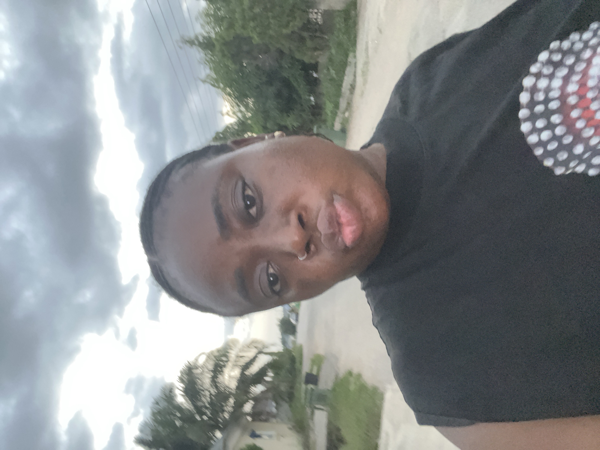 A close-up, off-kilter selfie shows a dark-skinned person with a nose ring and short, dark hair. They wear a black shirt with a pattern of white circles with red centers. The background is blurred, featuring a cloudy sky, trees, and what appears to be a street with buildings. The location is likely outdoors, possibly in a residential area. There are no definitive indicators placing the location in Abuja, Nigeria. Further evidence would be needed to confirm the location.