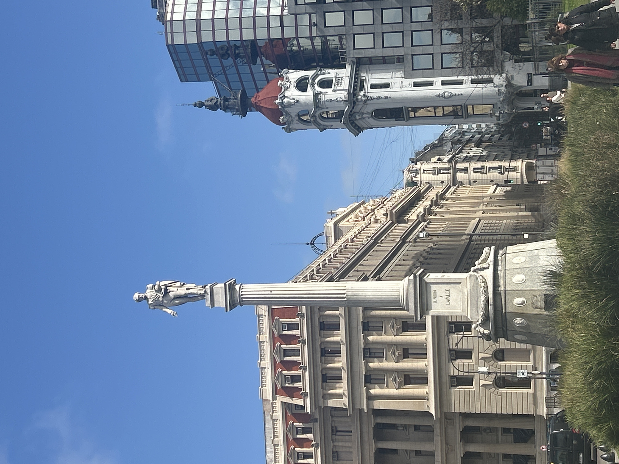 This image captures a street scene in Buenos Aires, Argentina, featuring a prominent monument and several buildings. The monument, a tall, fluted column topped with a statue of a man in military attire, stands in the foreground. The base of the monument is inscribed with "EL PUEBLO A LAVALLE". Behind the monument, a grand, classical-style building with red-tiled roofs and numerous windows dominates the left side of the frame. To the right of this, a distinctive white tower with a red-domed roof and ornate architectural details rises, dwarfed by a modern, glass-facaded skyscraper. The street is lined with buildings of varying architectural styles, suggesting an urban environment with a mix of historical and contemporary structures. A grassy median with tall, dry grass separates the monument from the sidewalk. Two women and a man are visible walking on the sidewalk in the lower right portion of the image. The sky is clear and blue, indicating fair weather. A black car is parked on the street in the lower left corner. Power lines are visible stretching across the scene.
