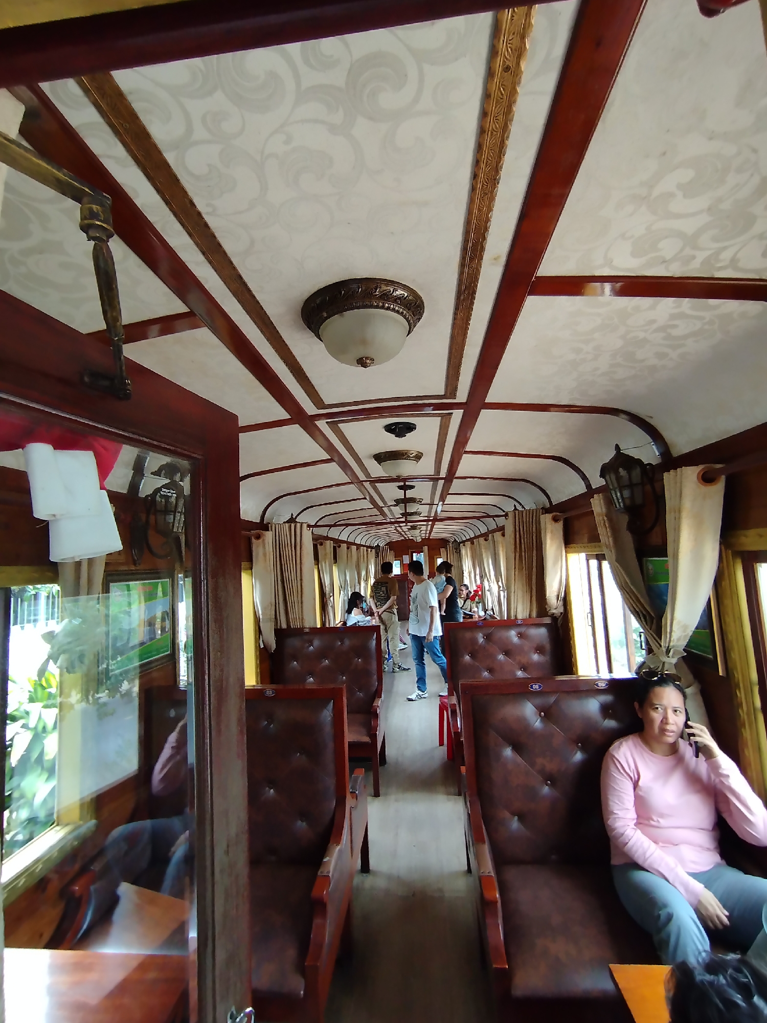 The image shows the interior of a vintage-style train carriage, likely a stationary tourist attraction, located in Lộc Quý, Vietnam. The carriage features polished dark wood framing, patterned ceiling panels, and ornate lighting fixtures. Rows of brown leather-tufted seats are arranged along the length of the carriage. Windows draped with light-colored curtains provide natural light. Several people are visible: a woman seated on the right side of the frame is talking on a cell phone, while other people are standing or walking further down the aisle of the train.
