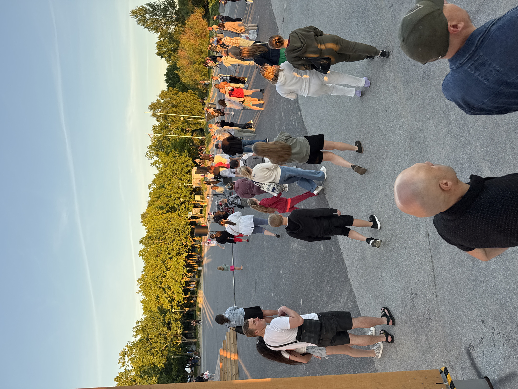 The image shows an outdoor gathering of people in a paved public space with trees in the background. The scene appears to be in Tallinn, Estonia, given the presence of a stage setup and what looks like a band on a platform, suggesting a concert or festival. The sky is clear and blue, indicating fair weather, and the sun is low, casting long shadows, implying it is late afternoon or early evening.
In the foreground, several individuals are visible from behind, walking away from the camera or looking in different directions. One man with a shaved head is in the lower center, wearing a black shirt, and another man wearing a denim jacket and a green baseball cap is to his right.
Further into the scene, a group of people are mingling. There are individuals of various ages dressed in casual clothing, including t-shirts, shorts, skirts, and pants. Some people are standing together, while others are walking. A young girl in a pink skirt stands out.
In the middle ground, a more organized setup is apparent. A stage with steps is present, and a band with drums and other instruments is visible. Speakers are set up on the ground. Several people are gathered around the stage area, seemingly watching or participating in an event.
The background is dominated by a line of lush green trees, characteristic of a park or a well-landscaped urban area. The overall atmosphere suggests a relaxed, community event taking place in a city park or outdoor venue.