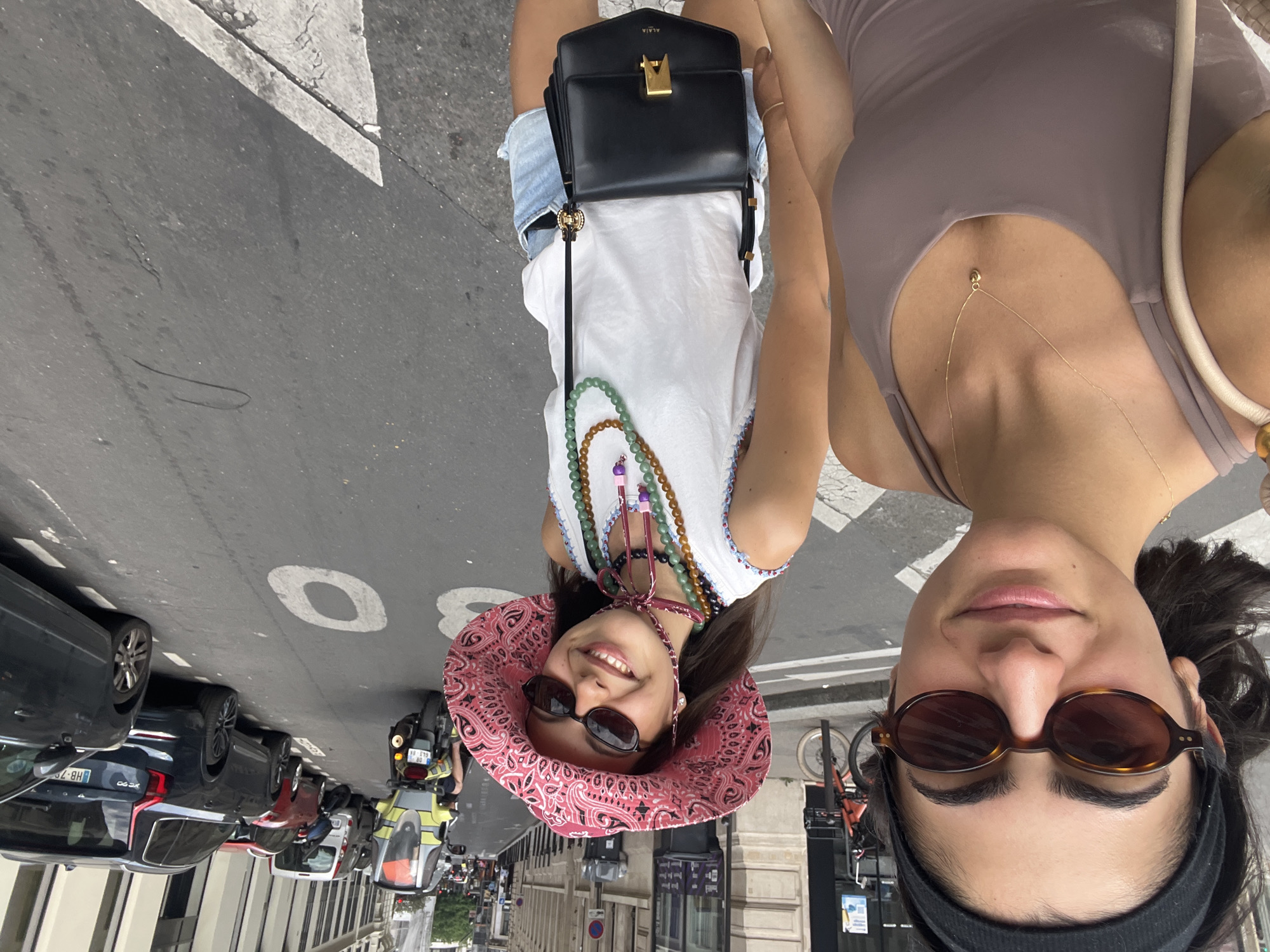 Two women are walking down a Parisian street. The woman in the foreground is wearing a dark headband, oval-shaped sunglasses, and a taupe tank top with a delicate gold necklace. The woman in the background is wearing a pink bandana-print bucket hat, dark sunglasses, a white tank top with colorful trim, and denim shorts. She is also wearing multiple beaded necklaces and carrying a black crossbody bag with a gold clasp. Several cars and a scooter are parked along the street. The street has white markings, including a directional arrow painted on the asphalt. The buildings on either side of the street appear to be typical Parisian architecture, with windows and awnings visible.