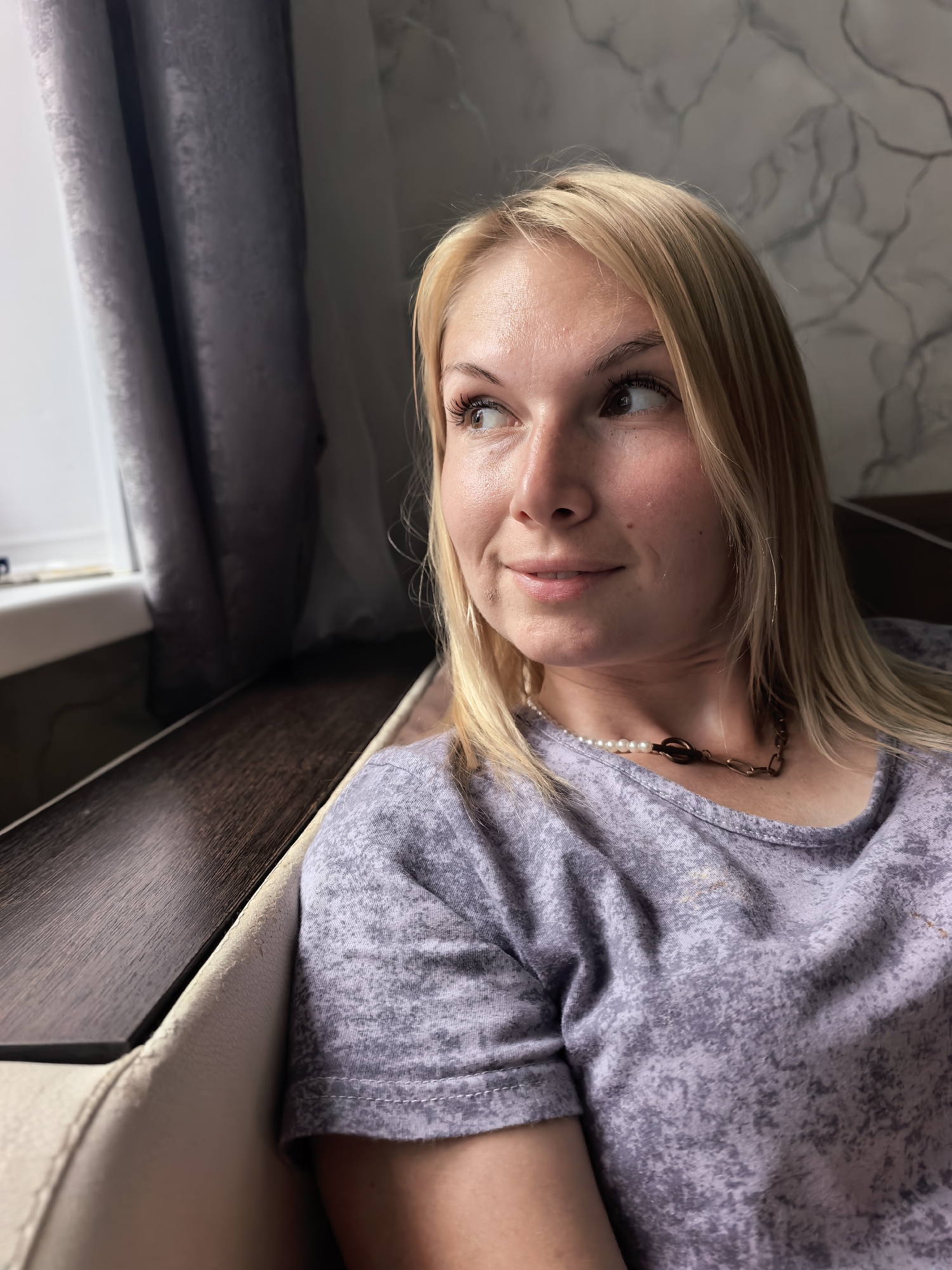 This media file is a close-up, medium shot of a young Caucasian woman with blonde hair, seated indoors. She is looking to her left, away from the camera, with a slight smile and a curious expression. Her eyes are brown, and she is wearing makeup, including mascara and eyeliner. She has a necklace with pearls and a metal link chain. Her shirt is a light purple color with a mottled pattern.
She appears to be sitting on a couch or bench with a dark wood surface and a light-colored cushioned seat. To her left, there is a window with a gray curtain partially drawn. The background wall has a marbled pattern. The lighting suggests it is daytime, possibly afternoon, with natural light coming from the window. The overall mood is relaxed and contemplative. There is no visible text in the image. The location context is Sterlitamak, Russia.