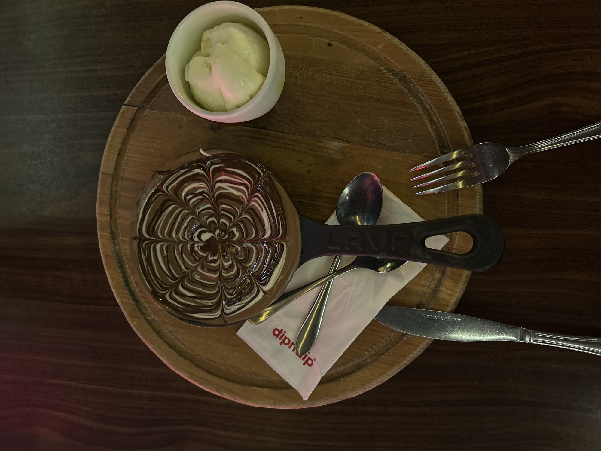 An overhead shot showcases a dessert platter on a dark wooden table, likely in an indoor setting. The main subject is a rich chocolate dessert served in a small, dark pan-like dish, featuring an intricate swirl pattern created with dark and white chocolate. The pan has "LAVA" embossed on its handle.
Accompanying the dessert, to its upper left, is a small white bowl holding scoops of light-colored ice cream, possibly vanilla. All items are presented on a circular, light-brown wooden serving board.
Cutlery includes a silver fork positioned to the upper right. Beneath the dessert, a folded white napkin bears the brand name "dipndip" in red text. Two spoons, one silver and one with a darker finish, rest on this napkin, while a silver knife is placed below it. The scene captures a prepared dessert, characteristic of a cafe or dessert lounge, ready for consumption. The lighting is subdued, suggesting an evening or ambient atmosphere. The context provided indicates the location is Kuala Lumpur, Malaysia.