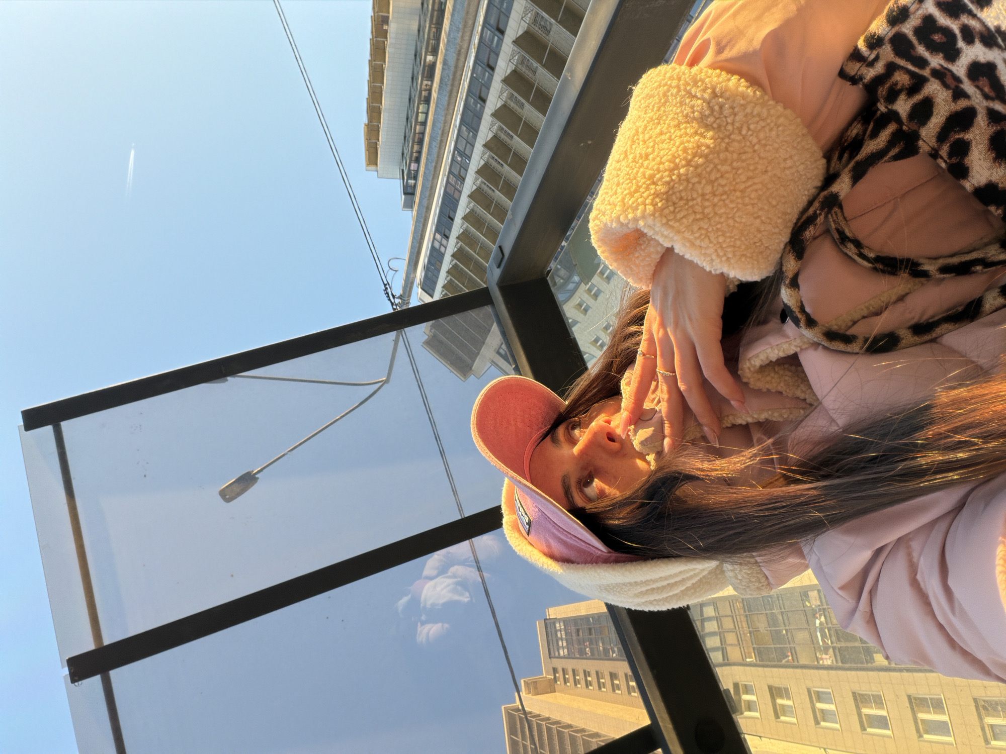 A low-angle shot of a woman in a pink baseball cap, a pink coat with a sherpa-like collar and cuffs, and a leopard-print bag. She has brown hair and is wearing rings. Her hands are near her face. The photo appears to be taken from a glass-enclosed structure, possibly a bus stop or an observation point, reflecting the cityscape. A high-rise building is in the upper background, while another building is visible in the reflection of the glass enclosure. A vapor trail is in the blue sky. The specific location in Saint Petersburg, Russia, cannot be determined from the provided image.