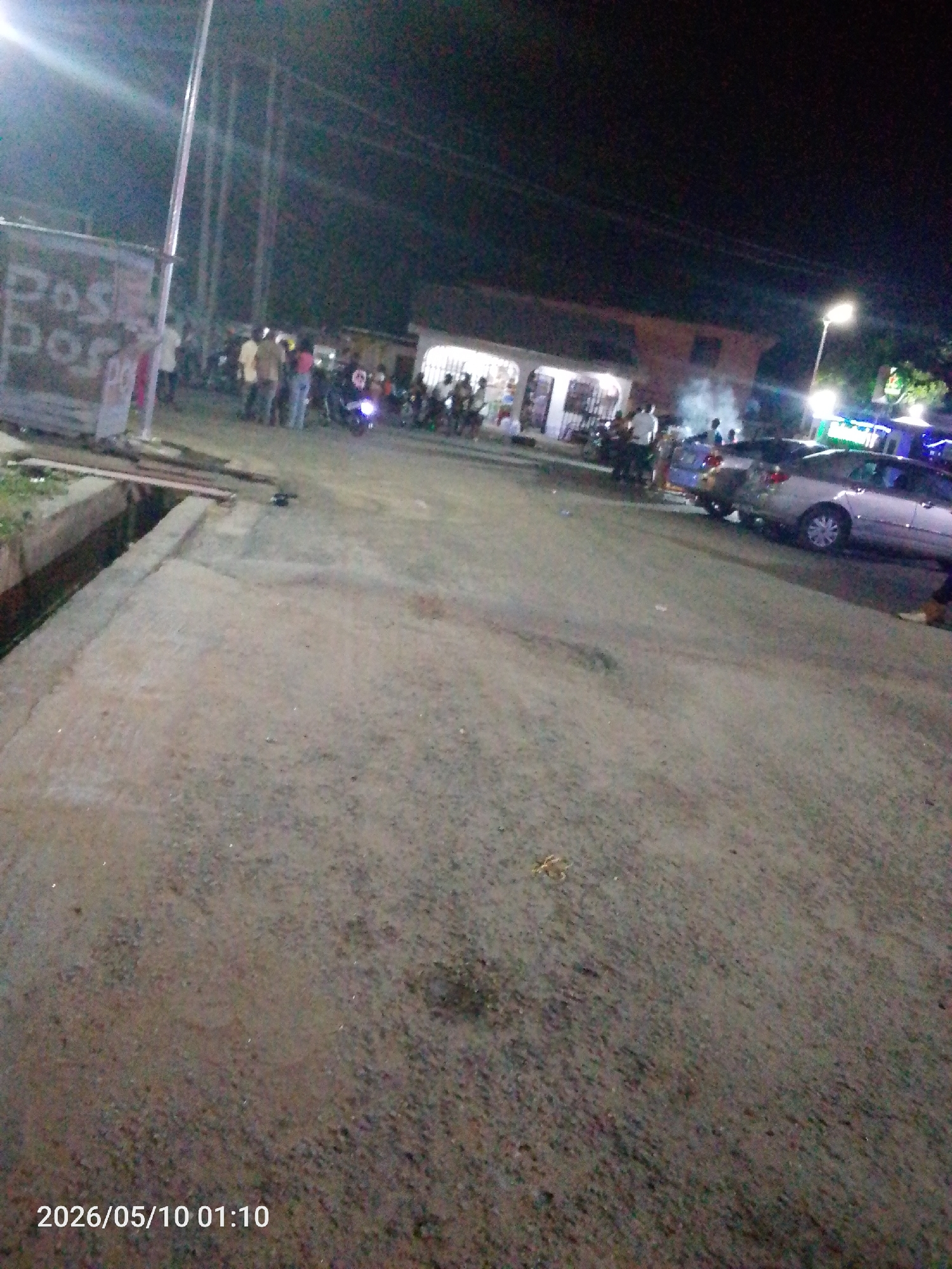 The image shows a street scene in Lokoja, Nigeria, at night. Several people are gathered near a building with a lighted storefront. A motorcycle is parked among the group. Cars are parked along the side of the street. Overhead lighting illuminates the scene. A concrete gutter runs along the edge of the road. The ground is paved. The date stamp indicates the photo was taken on May 10, 2026, at 01:10.