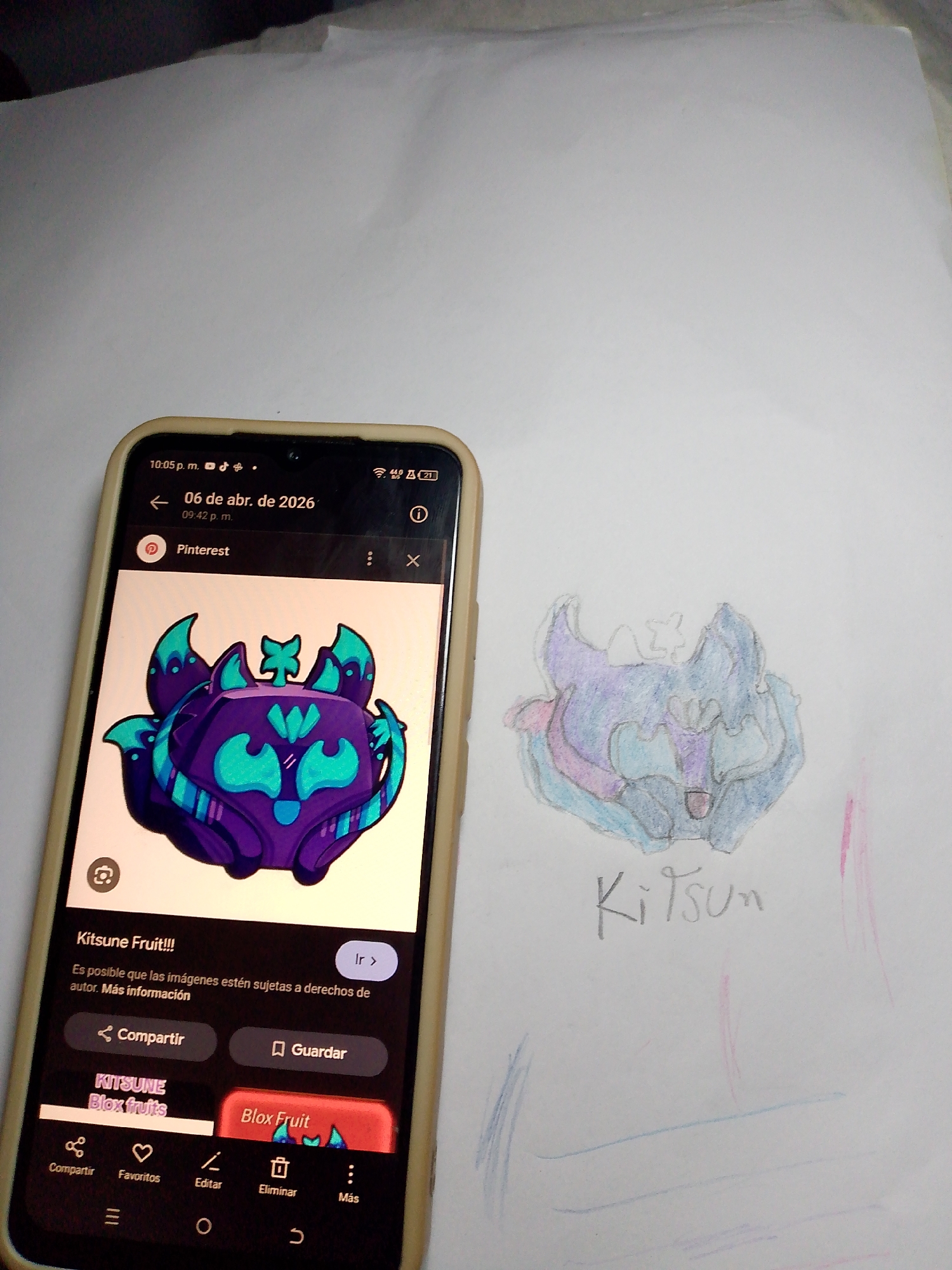 The image depicts a mobile phone lying on top of a sheet of white paper. The phone screen displays a Pinterest image of a cartoonish, stylized kitsune fruit, a purple and turquoise creature with fox-like features. Beside the phone, on the paper, is a roughly sketched, similarly designed kitsune fruit, rendered in light blue, purple, and pink. The word "Kitsun" is hand-written below the sketch. The location is Tinaquillo, Venezuela.