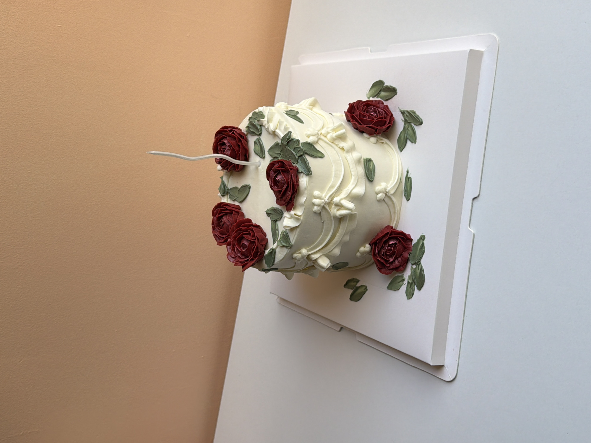 A white cake decorated with buttercream roses and leaves sits on a white cake board. The cake is topped with a single white candle. The background is a peach-colored wall. The scene is set on a white table. There is no indication of the cake's location in Tel Aviv, Israel.