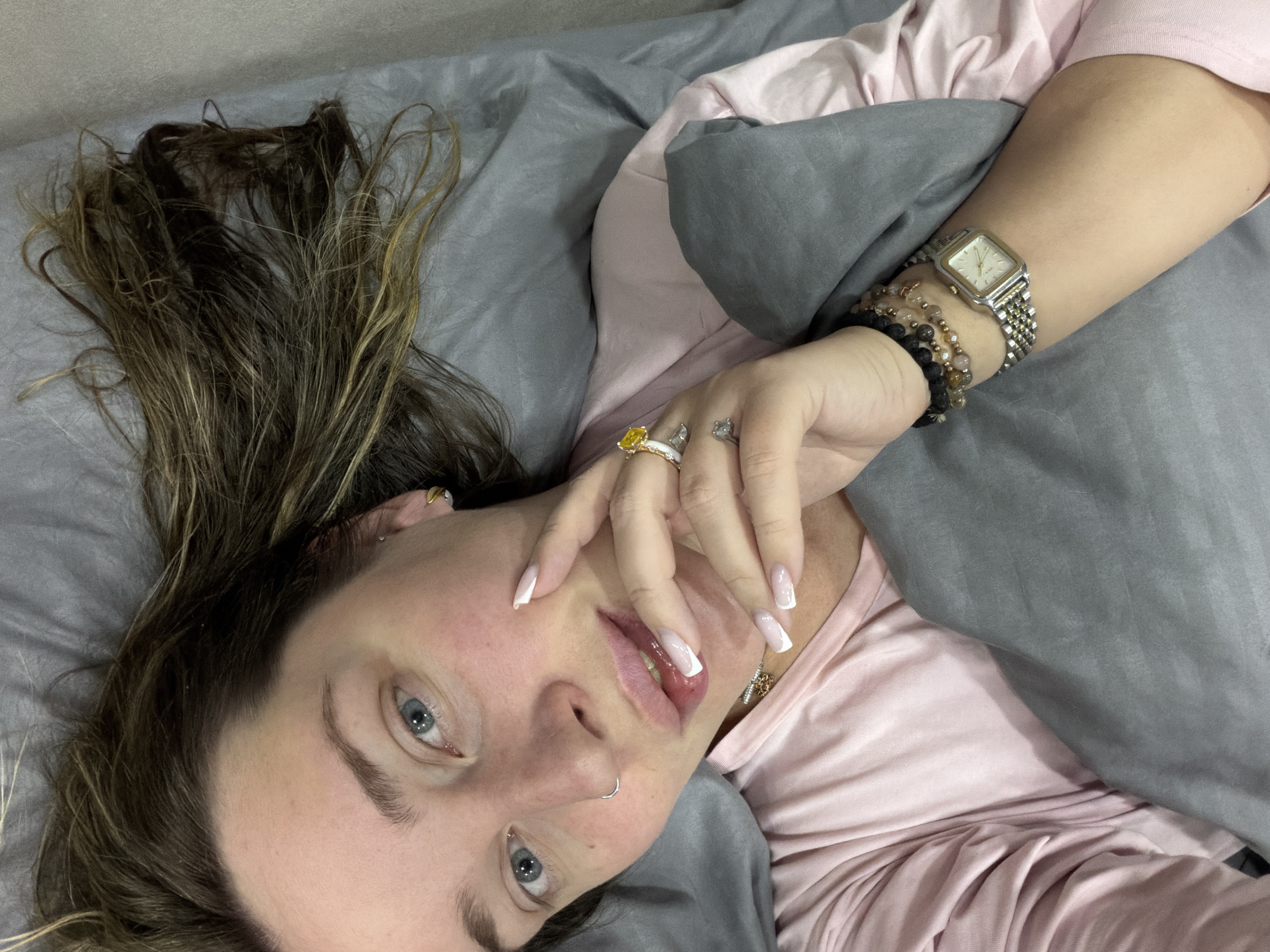 A close-up, overhead shot depicts a woman lying on a bed in Chaikovsky, Russia. She has fair skin, light blue eyes, and dark brown hair with lighter highlights, which is spread out around her head on the gray bedding. Her head is slightly tilted, and she gazes directly at the camera with a calm expression.
She is wearing a light pink top. Her right arm is bent, with her hand resting near her mouth. On her right hand, she wears several rings: a notable yellow gemstone ring on her ring finger, a smaller silver-toned ring adjacent to it, and a small, animal-shaped ring (resembling a cat) on her pinky finger. Her fingernails are long and styled with white tips. A small silver hoop nose ring is visible on her left nostril.
On her left wrist, she wears a square-faced analog watch with a two-tone (silver and gold) metal link band, featuring the brand "EXCEL" on its face. Alongside the watch are multiple beaded bracelets in varying shades of dark and light brown/amber. The bedding consists of a gray duvet cover and pillow, with a pink sheet or blanket layered beneath. The scene conveys a relaxed, indoor moment, likely in a bedroom.