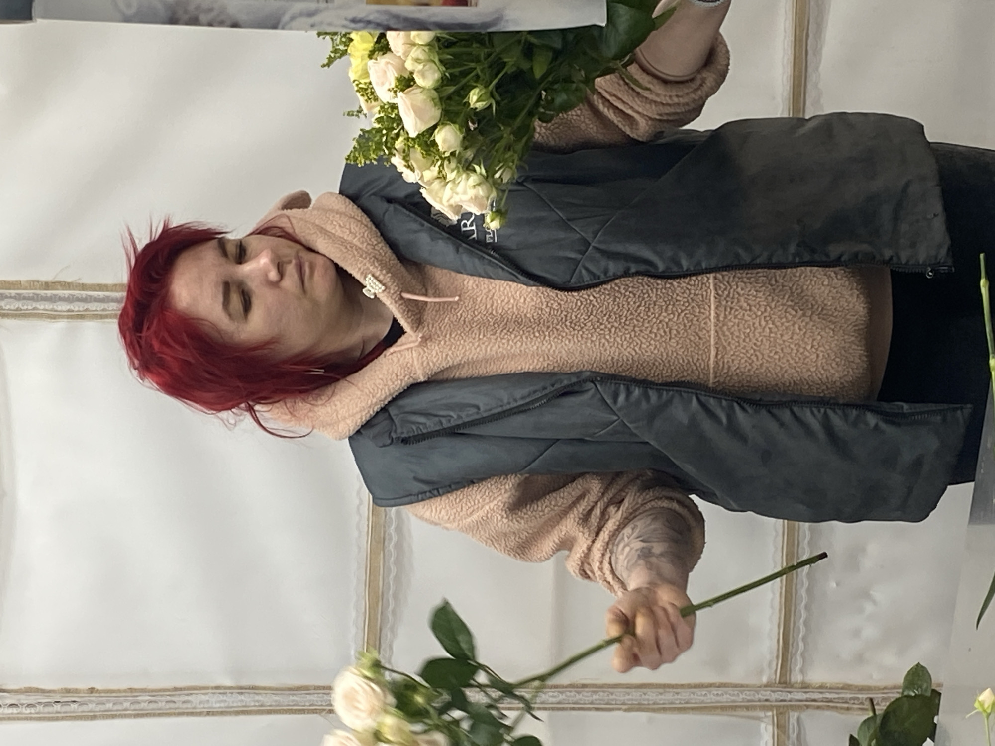 A woman with red hair is holding a bouquet of white roses. She is wearing a light brown, textured hoodie and a dark gray vest. The background appears to be a white wall with a decorative border. The location is Yekaterinburg, Russia.