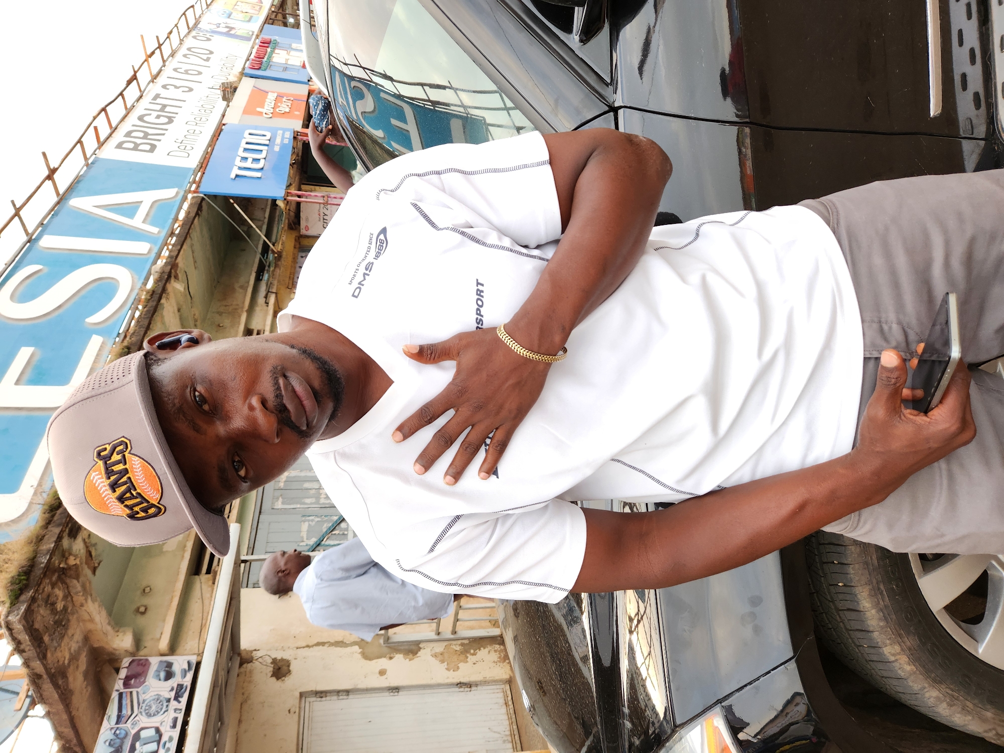 A dark-skinned man wearing a white t-shirt and a grey baseball cap with an orange and yellow "GIANTS" logo stands in front of a black SUV. The man has his right hand on his chest and is holding a dark-colored smartphone in his left hand. He is looking towards the camera with a neutral expression. Behind him, a faded building with a blue sign that reads "BRIGHT 3|6|20" and other businesses are visible. Another man in a light blue shirt is walking away in the background. The scene appears to be an outdoor market or commercial area.