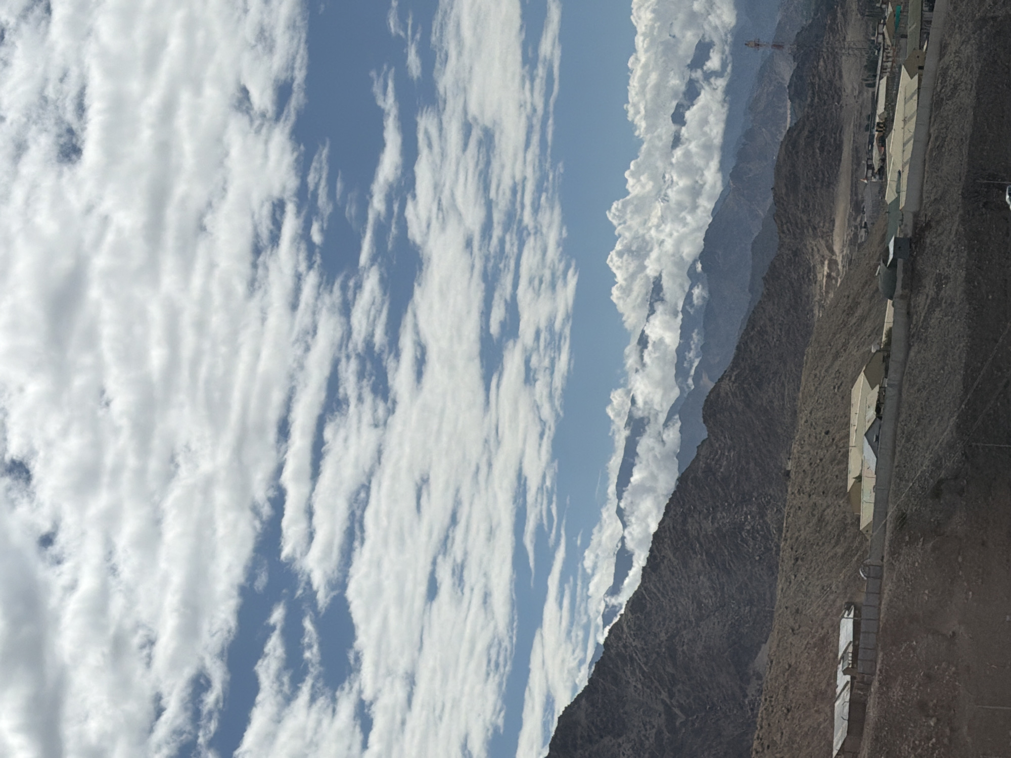 The image shows a mountainous landscape with a small settlement in the foreground. The settlement consists of several low-rise buildings with corrugated metal roofs, enclosed by a fence. The buildings appear to be industrial or commercial in nature. In the background, a series of rugged, rocky mountains rise, with some clouds clinging to their peaks. The sky is a vibrant blue, filled with scattered white clouds. A cell tower is visible on the right side of the image, suggesting some level of infrastructure in the area. The overall impression is one of a remote, arid, and elevated region.