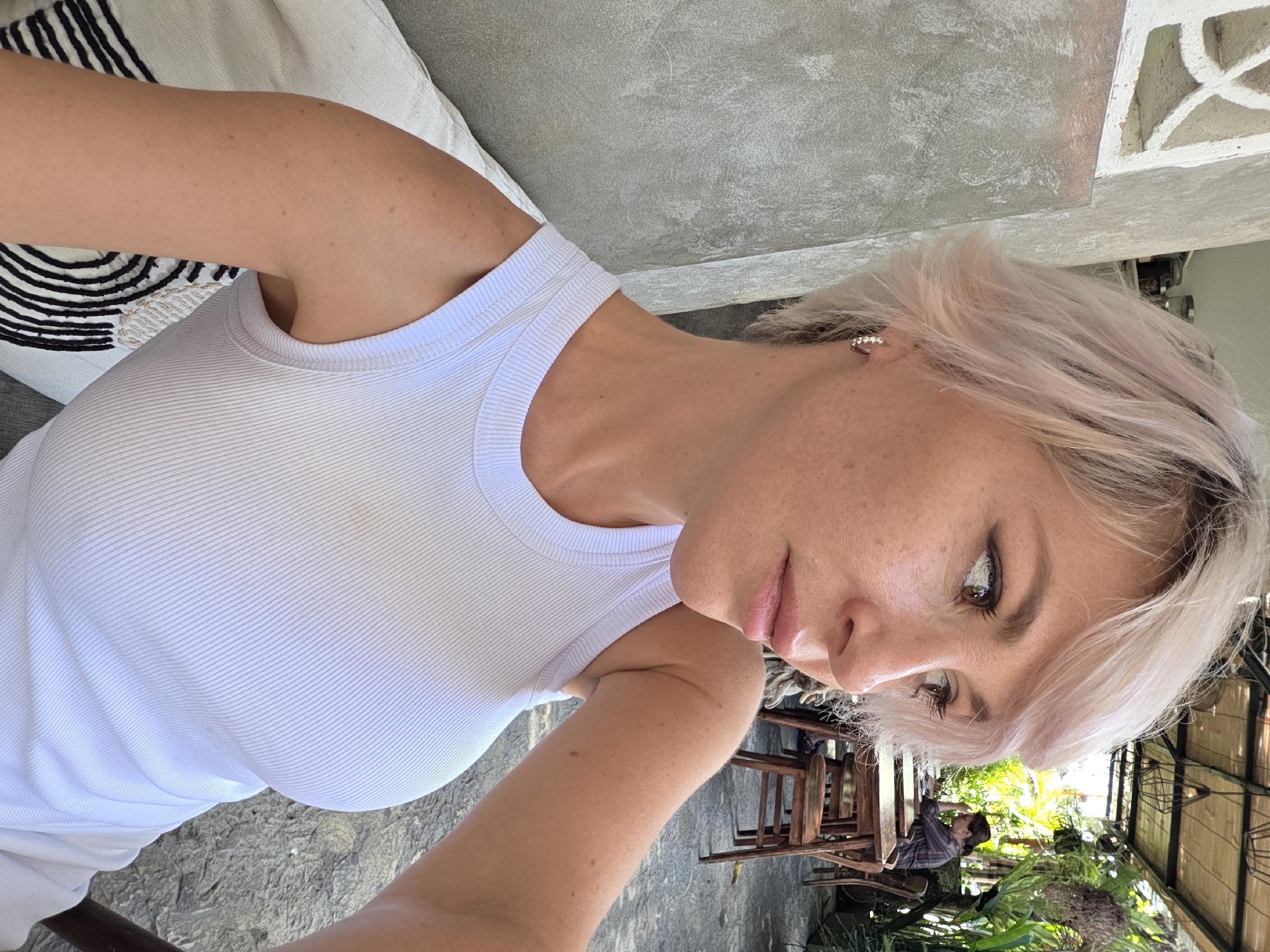 A light-skinned woman with short, light pink hair, wearing a white ribbed tank top and a small hoop earring, is seated at an outdoor cafe. She is looking to her right with her head slightly tilted. In the background, several wooden tables and chairs are visible, along with lush green foliage and a person seated at a table. The cafe appears to be located in North Kuta, Indonesia.