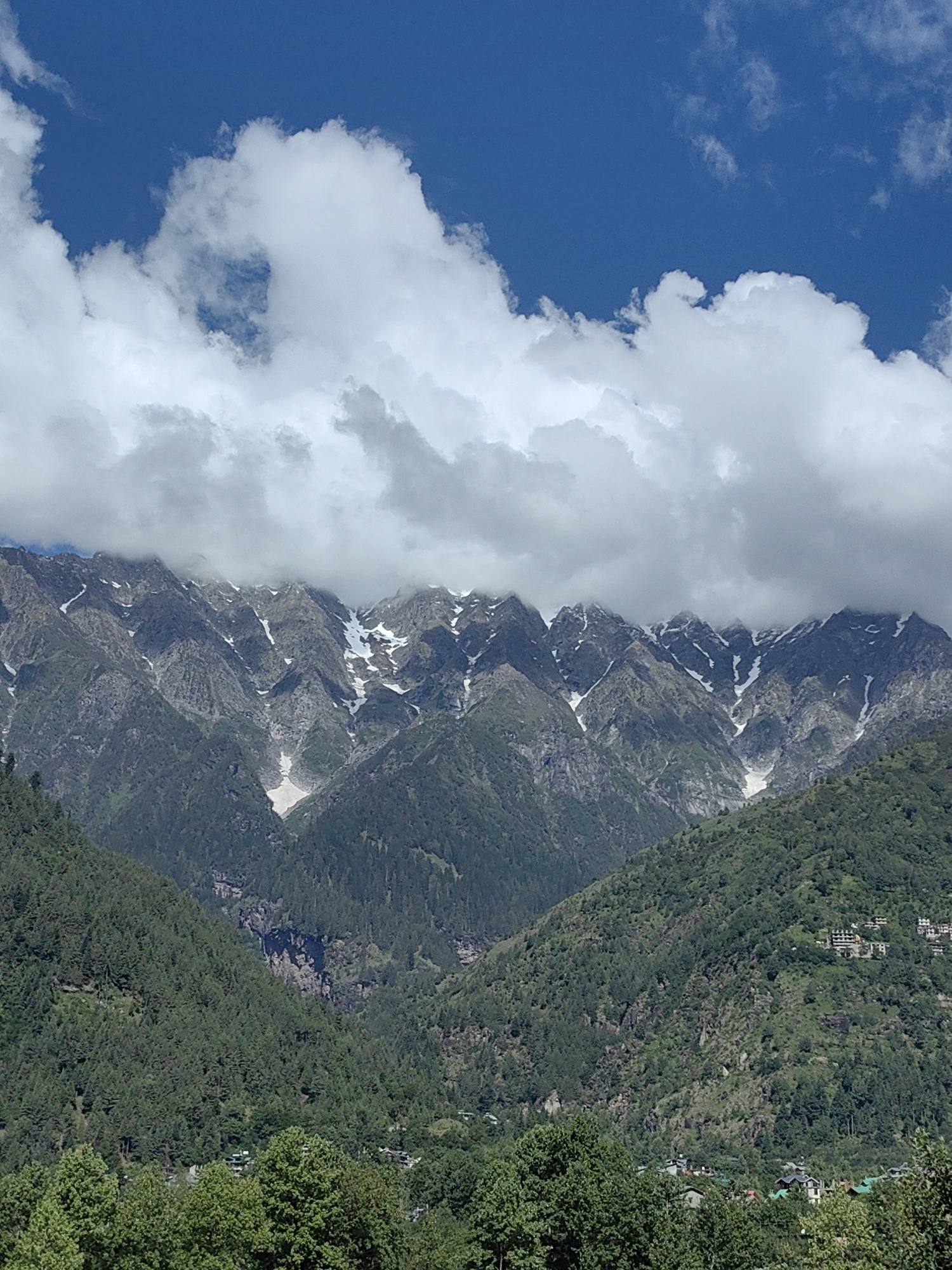 The image captures a majestic mountain range under a partly cloudy blue sky. Snow-capped peaks rise majestically in the background, their rugged slopes interspersed with patches of white snow. The mid-ground is dominated by lush green hillsides covered in dense pine forests. Scattered across the hillsides are small clusters of buildings, indicative of a settlement. The foreground features more dense green trees. The scene appears to be a scenic view of the mountains near Kulang, India, possibly taken from a higher vantage point. There are no people or animals visible in the image, and no specific actions or interactions are depicted. The environment is natural and serene, showcasing the beauty of the mountainous landscape.